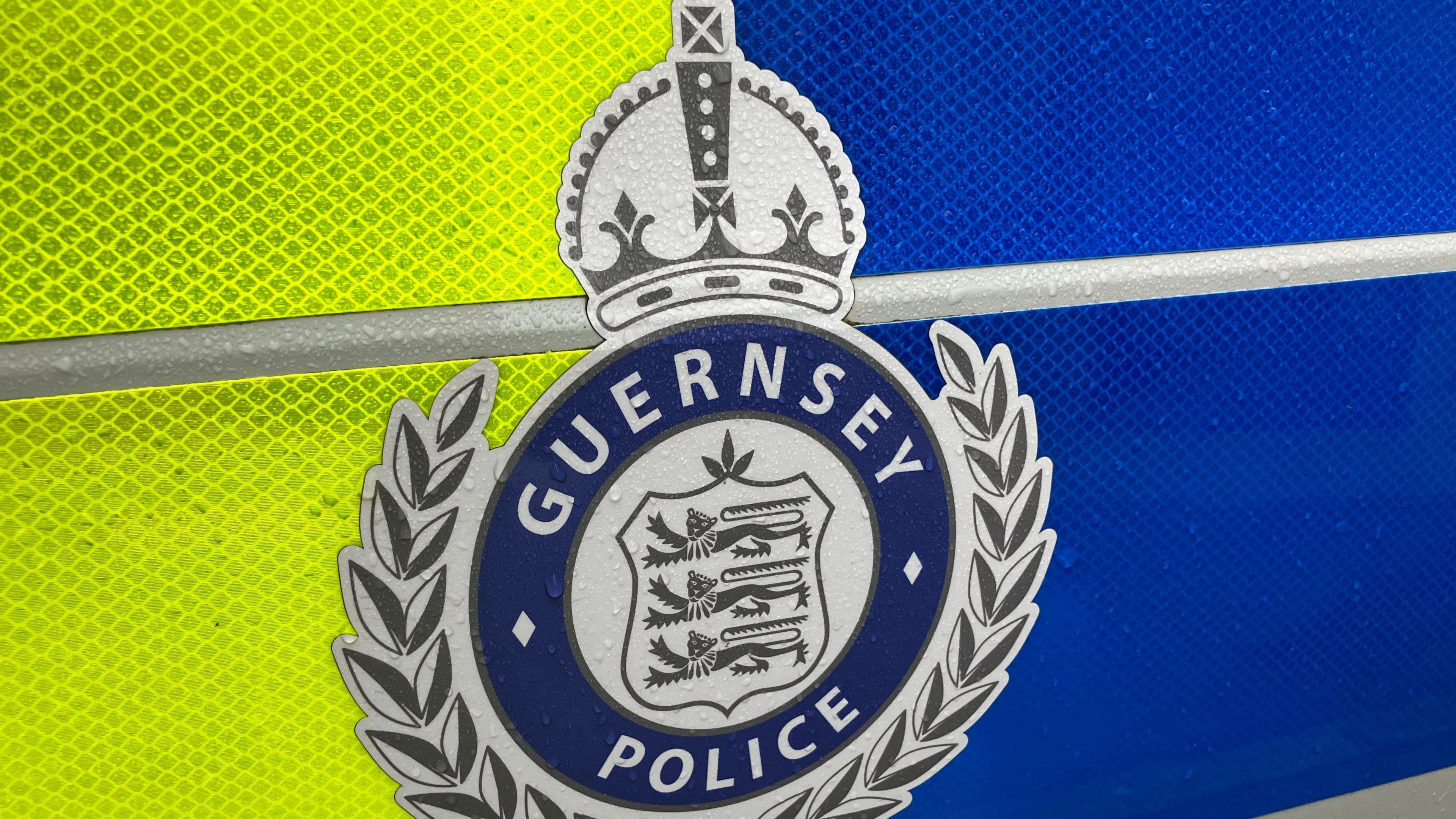 A coat of arms featuring three blue lions, with a laurel wreath and the words Guernsey Police surrounding them is on a fluorescent yellow and dark blue background.