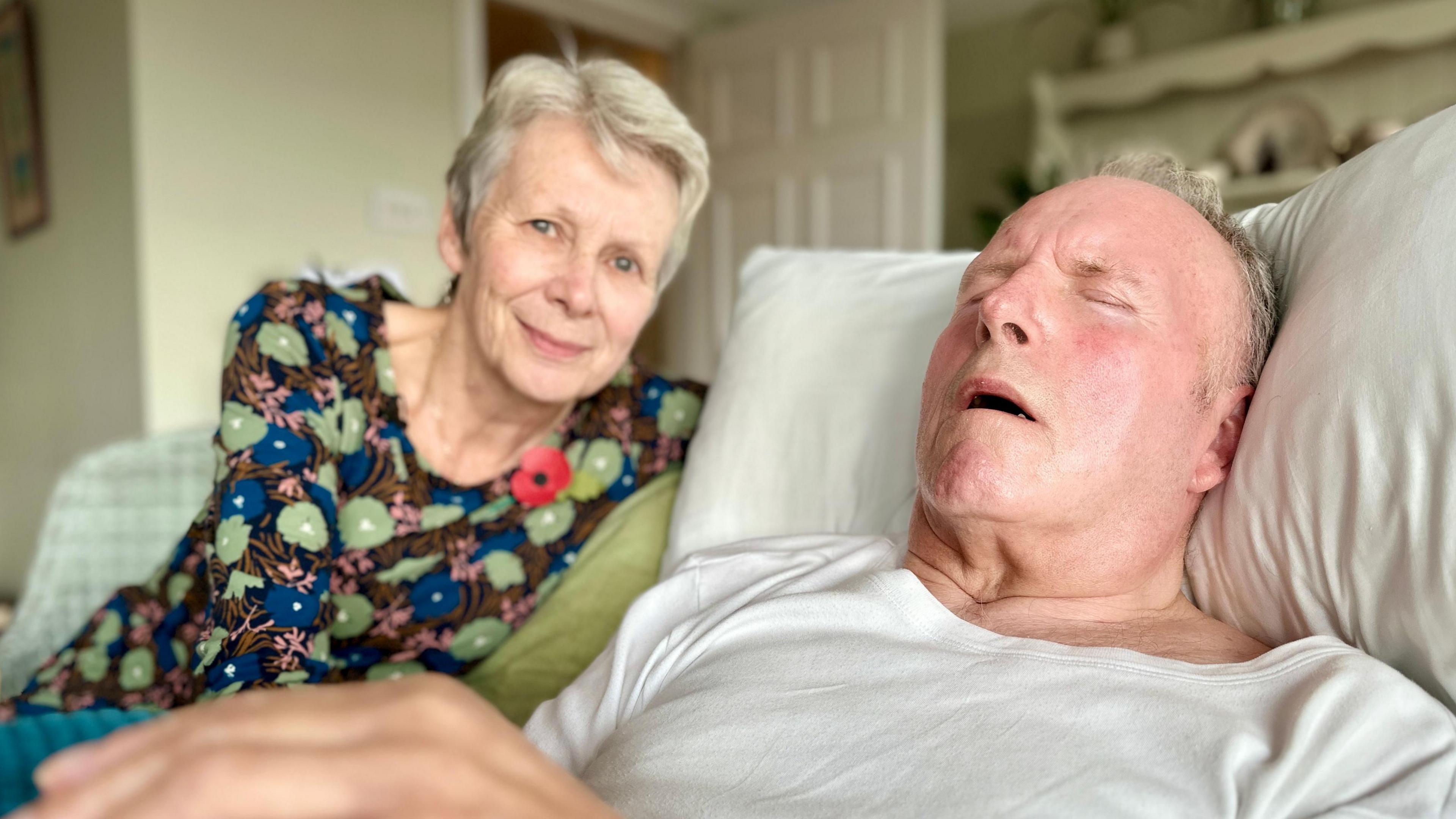 This is a head and shoulders picture of Anne Scowen and her husband Ray. Ray is lying on a bed with his head on a pillow in the foreground and has his eyes shut. Anne who has short grey hair and is wearing a floral dress, is sitting in a chair next to him, smiling. 