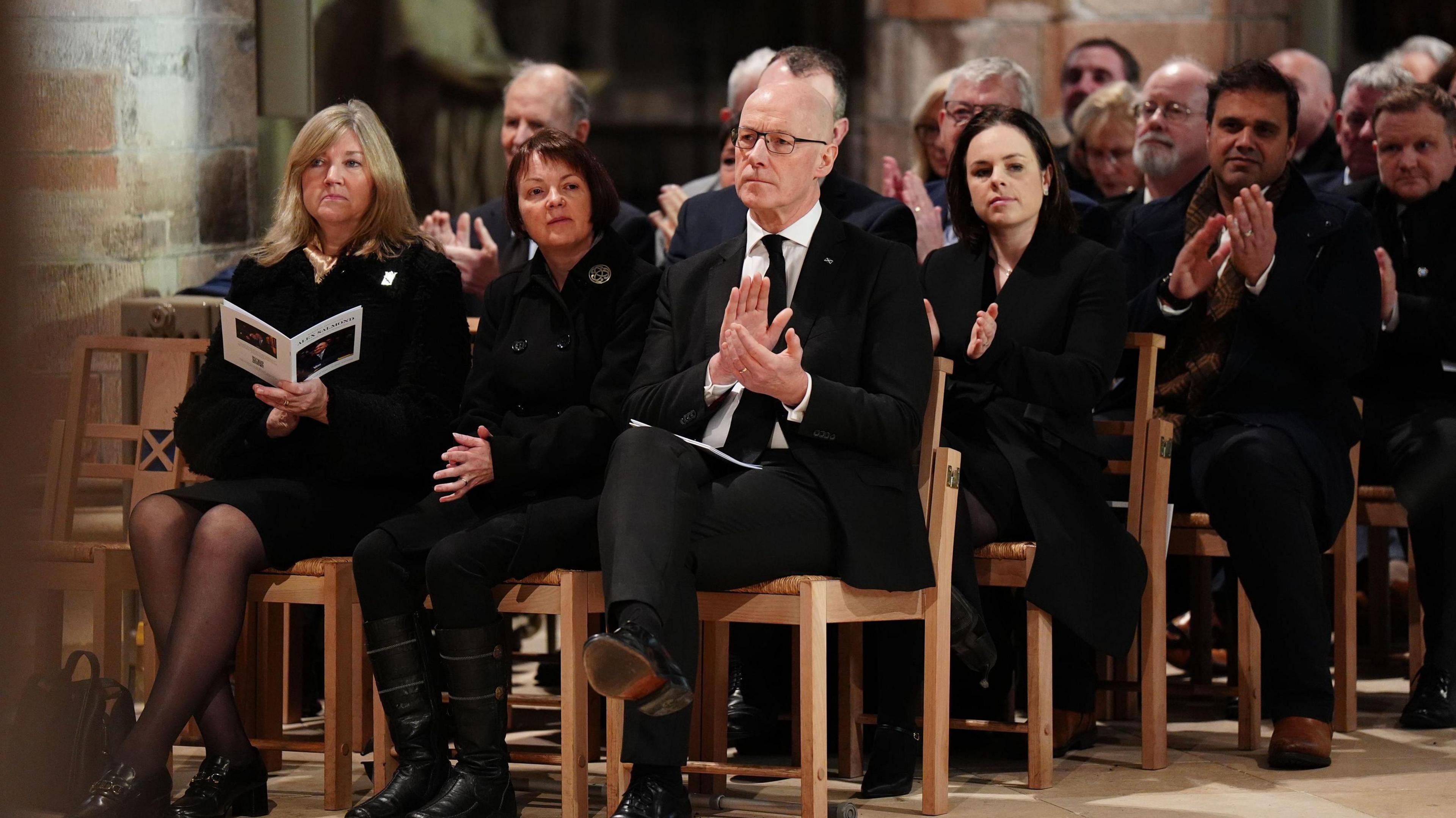 John swinney sits on a wodden seat with kate forbes behind him, and his wife and the presiding officer to his left. All are wearing black.