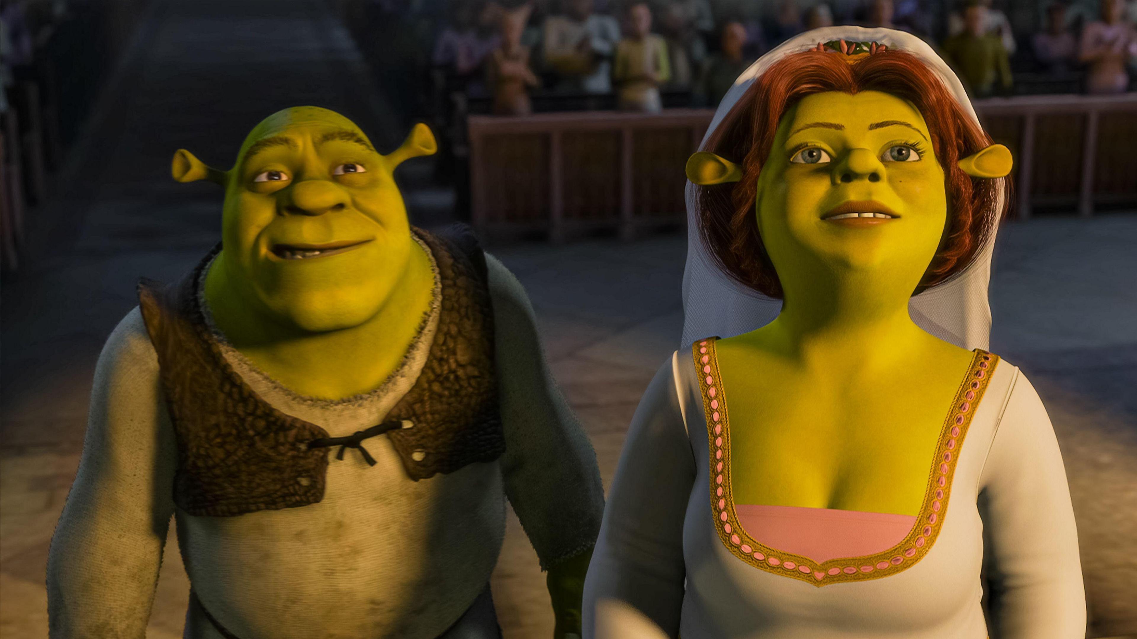Shrek and his wife Fiona stood side by side. She is wearing a wedding dress with a veil, she has ginger hair. Shrek is wearing a white long sleeved top with a small brown waistcoat.