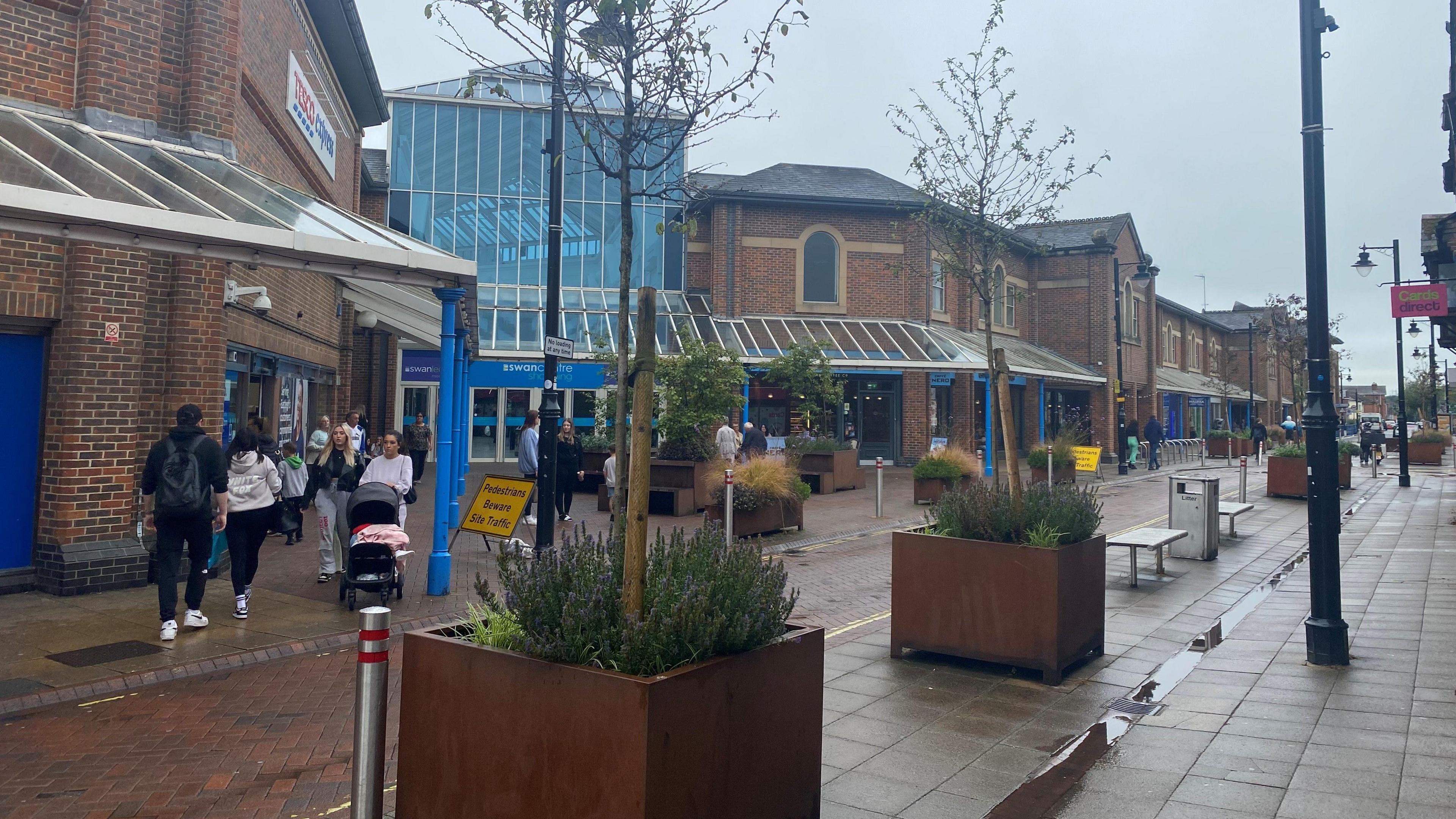 Eastleigh town centre, a built up street with people milling around. There are puddles on the floor. The street is lined with planters containing shrubs and trees.