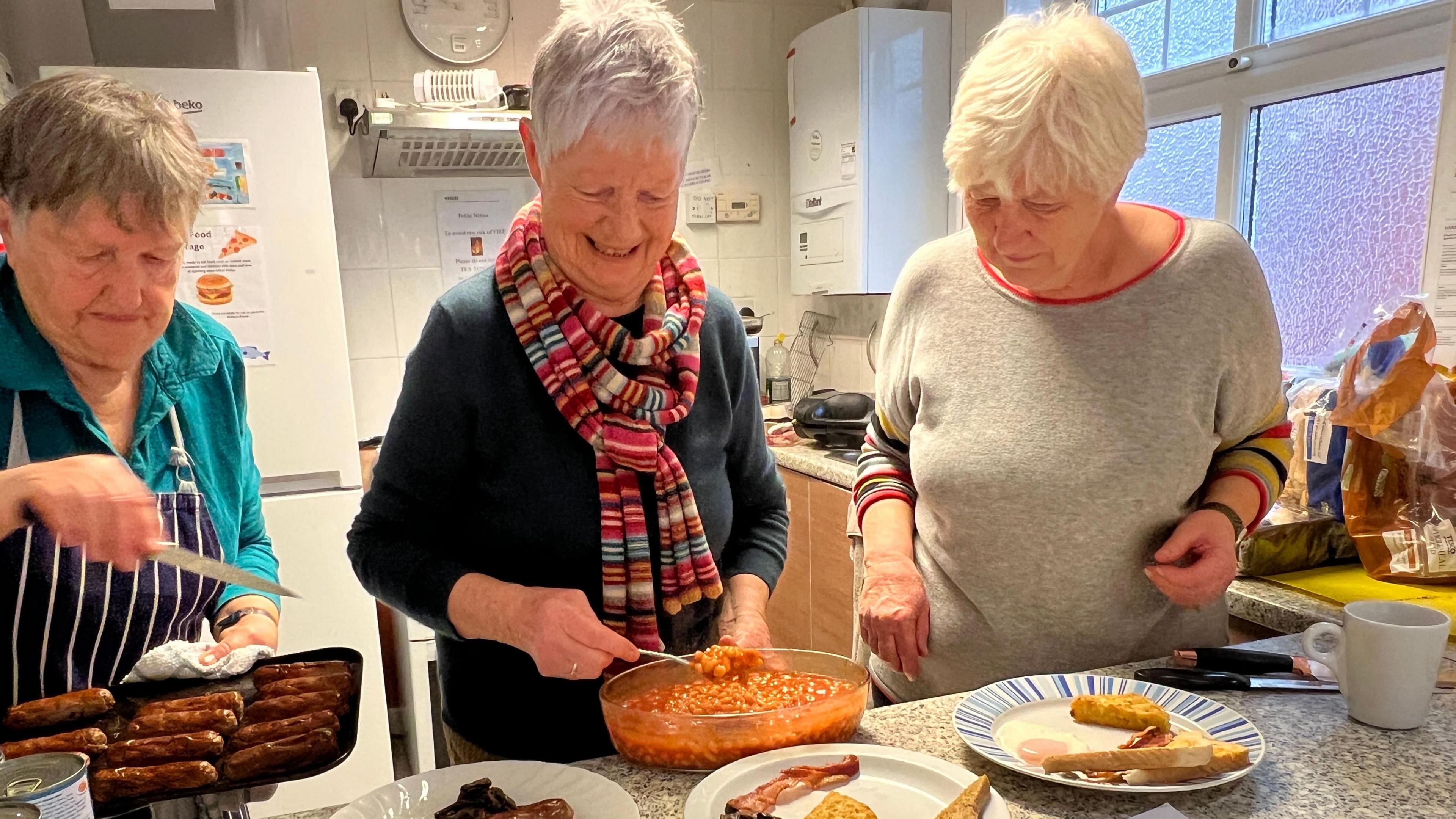 Three women stand in a row in a kitchen. The woman on the left holds a baking tray filled with cooked brown sausages while the woman in the middle spoons baked beans from an oval dish on to a plate that already has bacon and toast on it. 