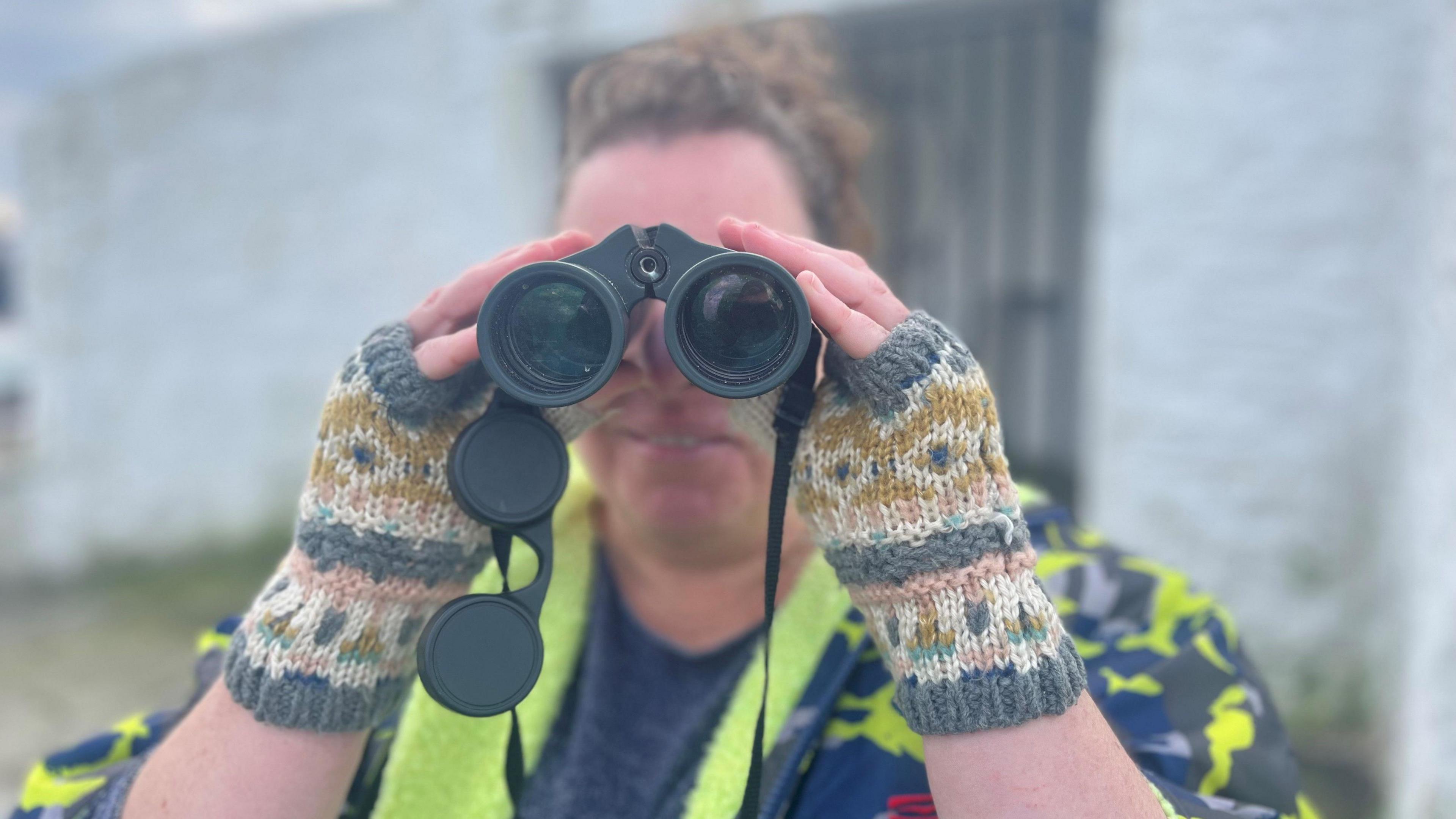 Angie Nash is looking at the camera through a pair of binoculars. She is smiling and  wearing a pair of woolly fingerless mittens and has her hair pinned up on her head.