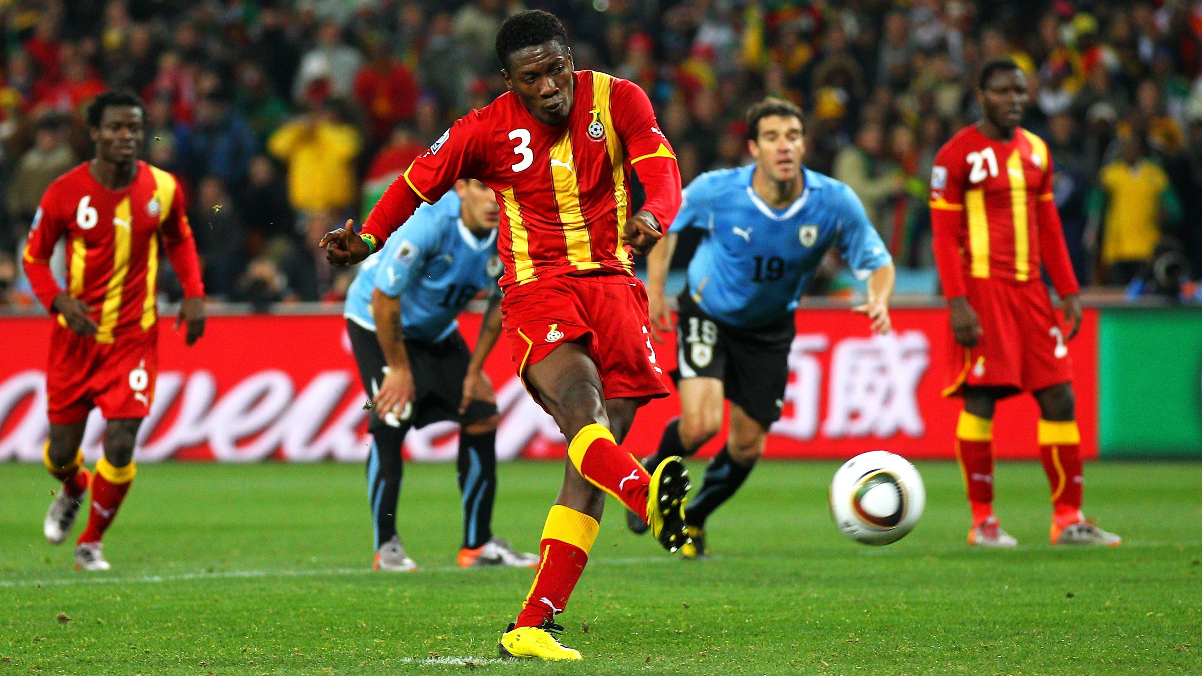 Ghana's Asamoah Gyan strikes his missed penalty in the quarter-final of the 2010 World Cup against Uruguay. Gyan is wearing red and gold socks, red shorts and a shirt made up of red and gold vertical stripes. A white number three is positioned in a patch of red on his right shoulder. He stands on his left leg while his right swings through his shot, with the blurry ball which he has just kicked seen rising away from the green turf. In the background, positioned on the edge of the penalty area, are two of Gyan's Ghana team-mates and two Uruguay defenders dressed in black socks and shorts and light blue jerseys. One of the defenders is beginning to run into the box while the other bends down with his hands on his knees