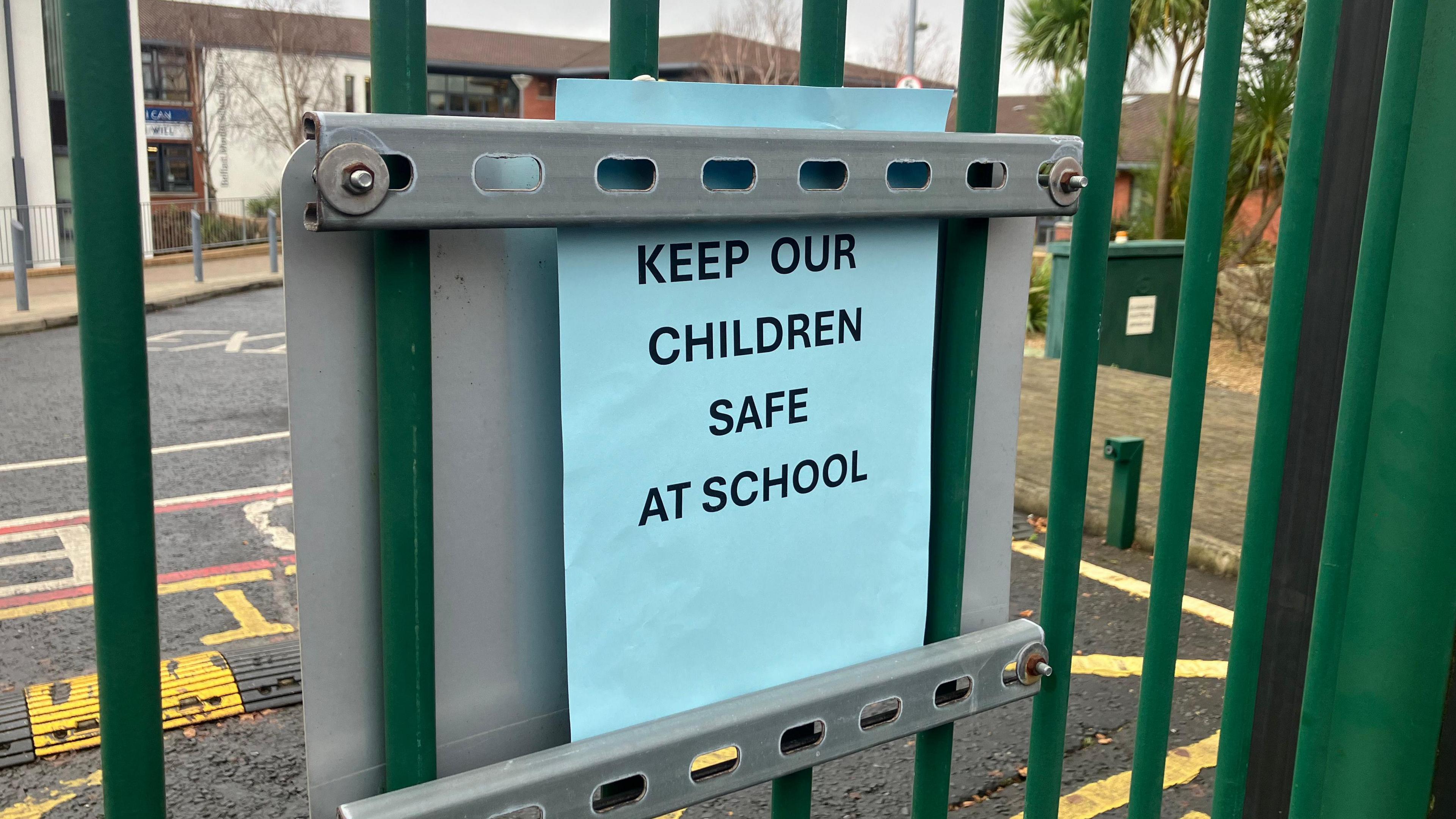 On the green metal gates there is a blue sign which reads 'Keep our children safe at school'.