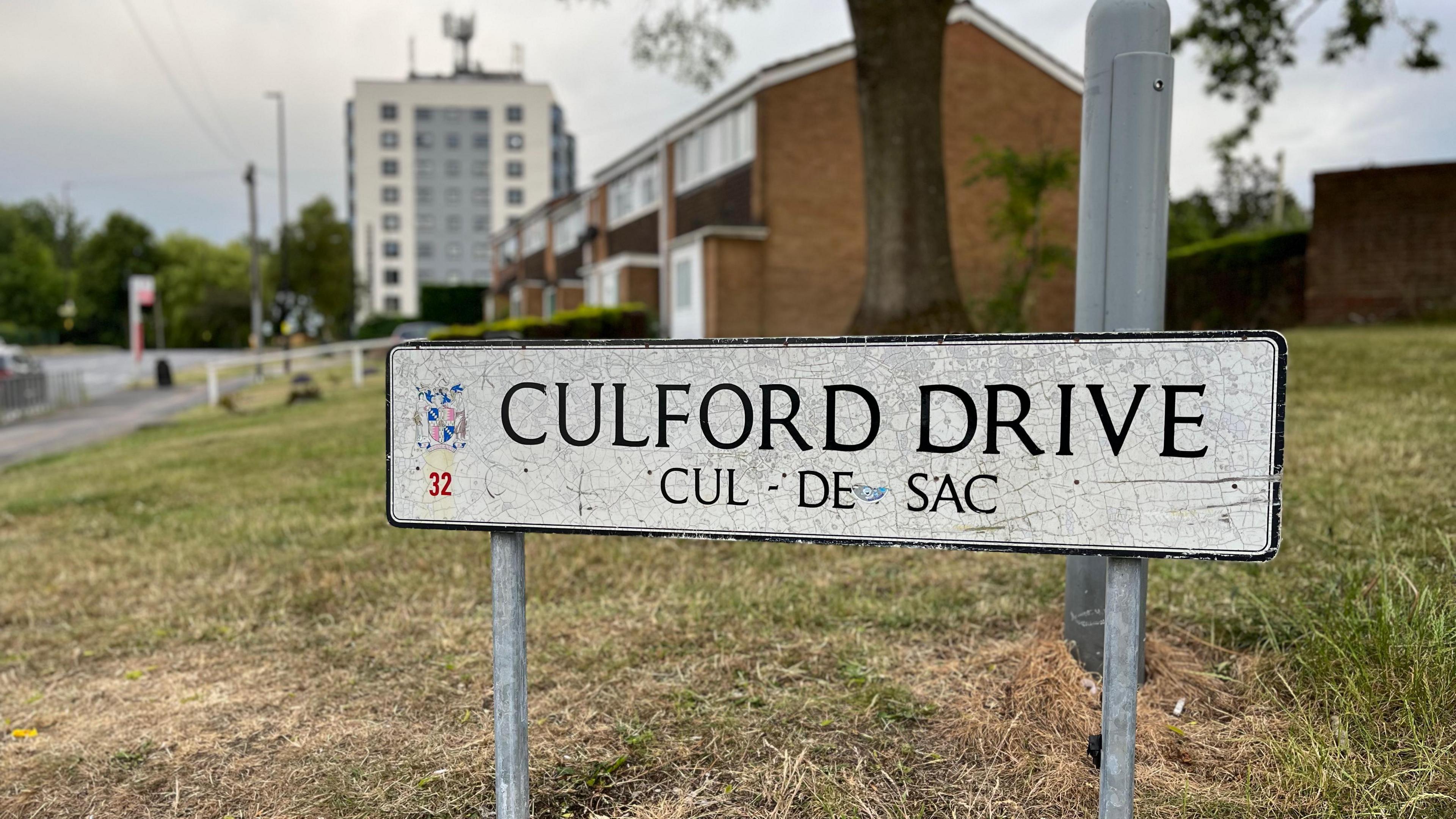 A street sign of Culford Drive, a cul-de-sac. It is old and scratched. The background is blurred but we can see some residential homes and a tower block behind it.