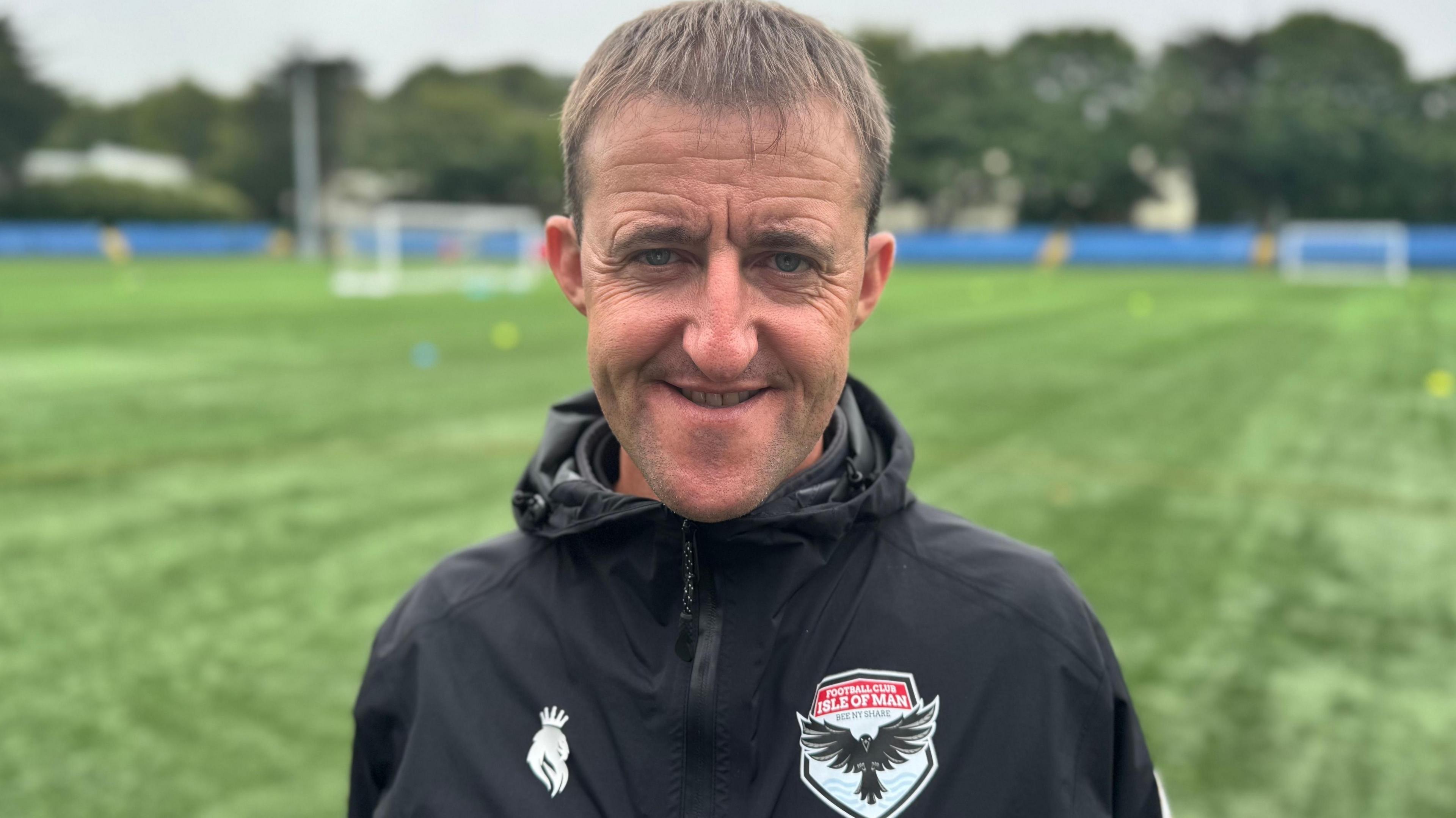 Lee Dixon wears a black rain jacket with the FC Isle of Man logo on the left side of his chest. He smiles. He has short fair hair and stands by a green football pitch.