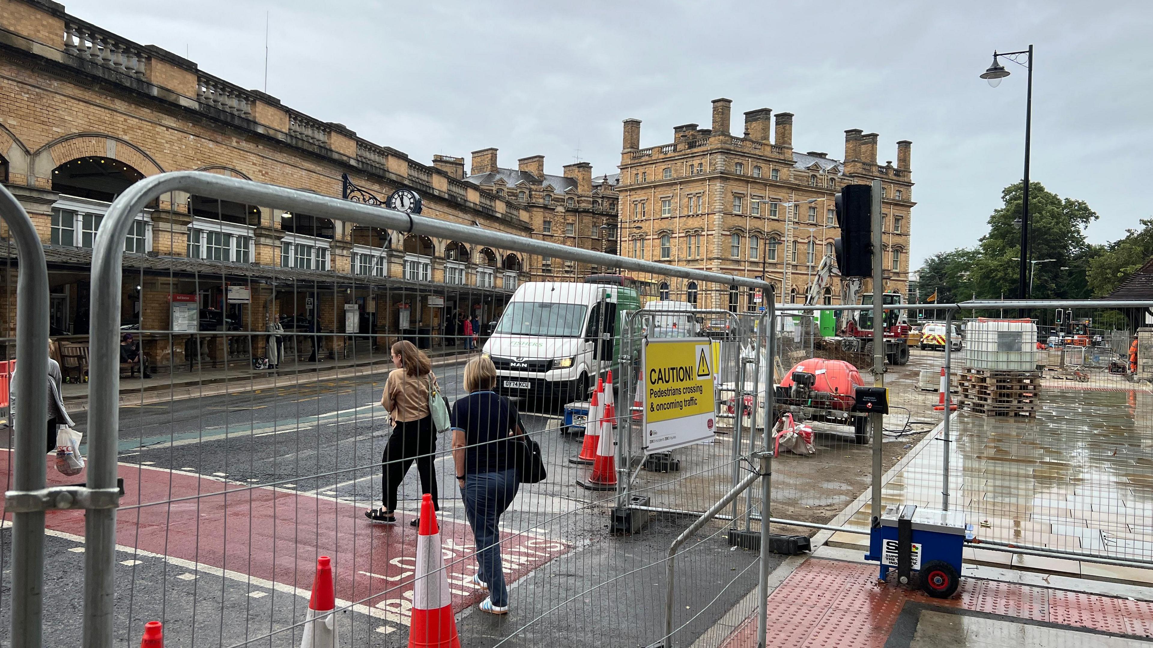 This is a picture of the ongoing work on York's Station Gateway scheme. Metal fences have closed off part of the area opposite the station entrance where new paving stones are being put in. People using the temporary pedestrian crossing are walking past traffic cones.