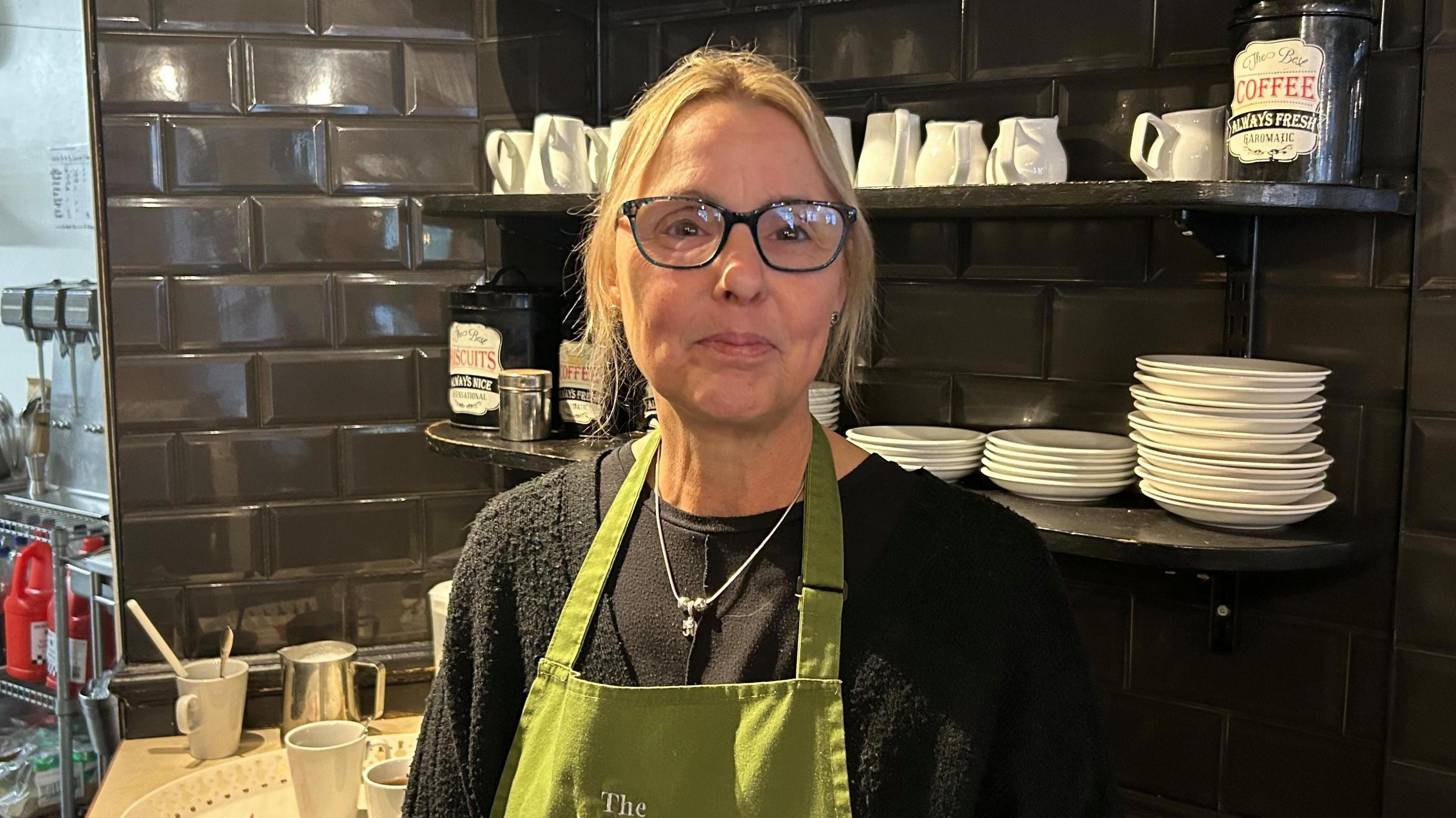 Sara Smith in her Lydney café standing front of shelves with plates and milk jugs. She has blonde hair, black-framed glasses and is wearing a green apron.