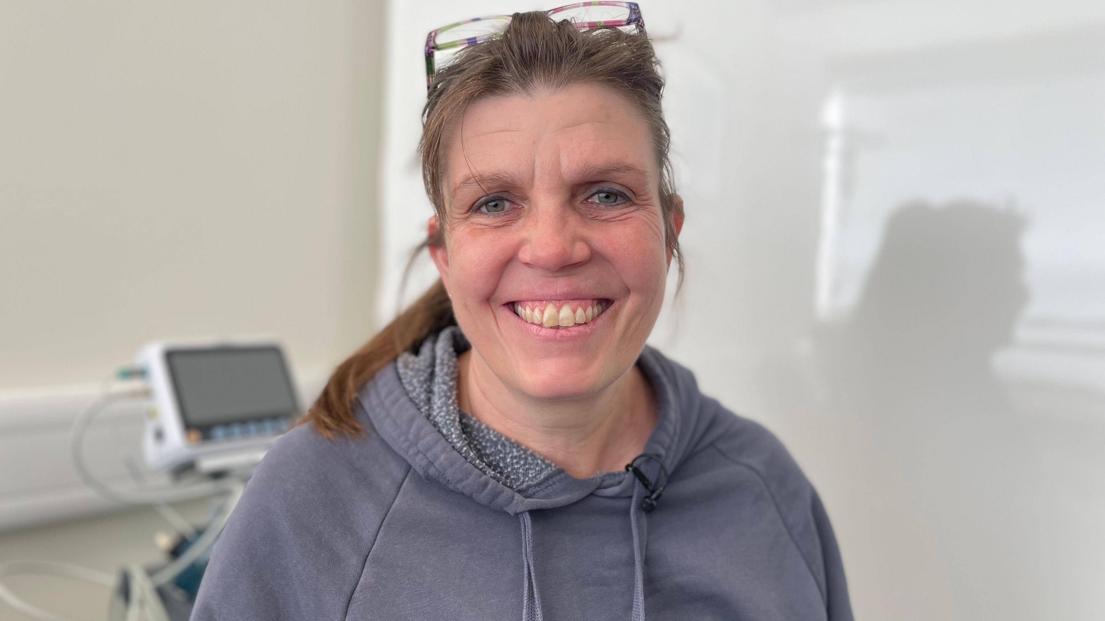 Kirsty Mitchell stands in the same hospital room as her husband, with a white background and medical equipment. Her brown hair is tied back and her glasses are on her head. She smiles and is wearing a grey hoodie.