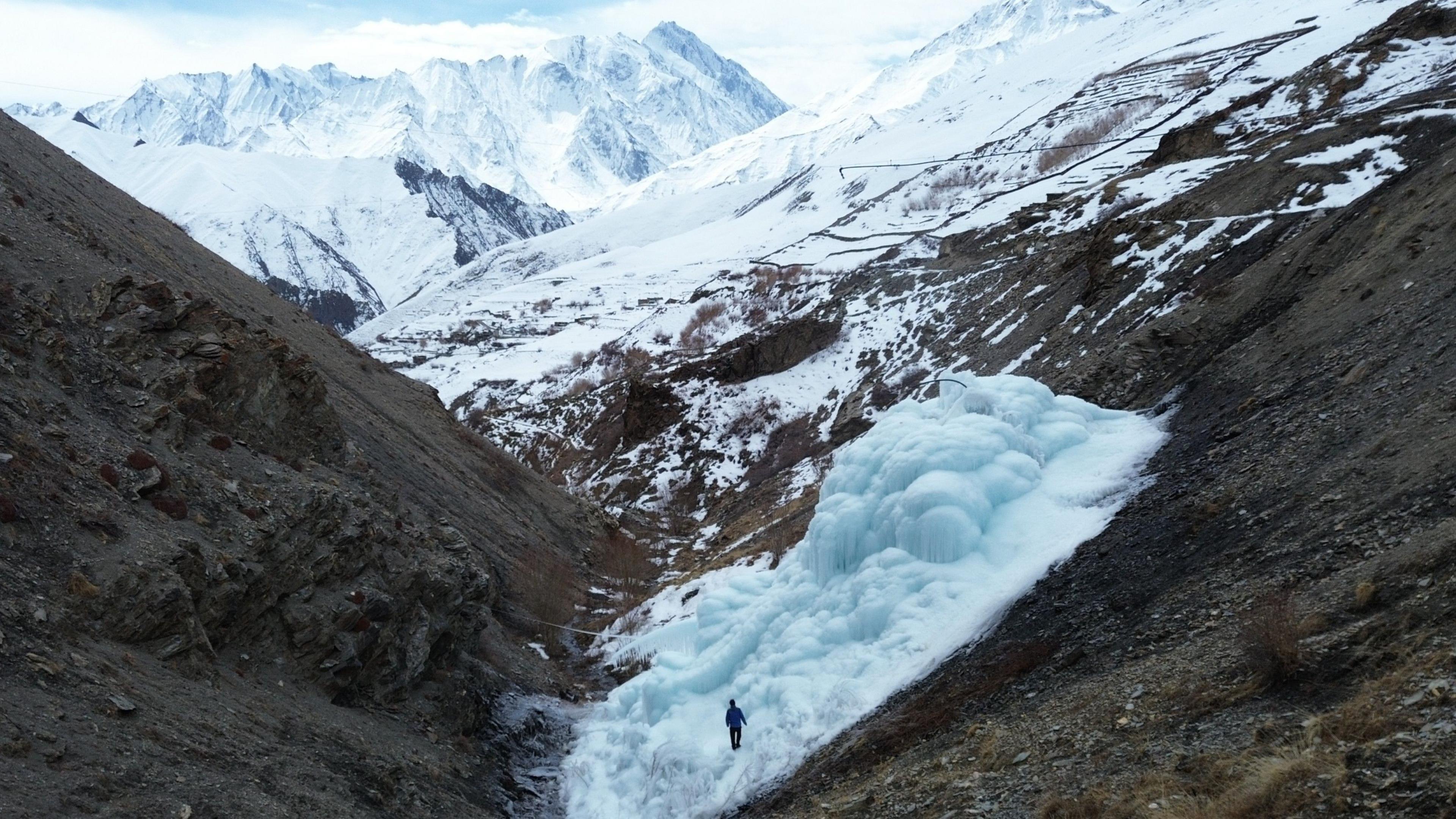 A man is dwarfed by an ice tower. In the distance are snow-covered mountains.