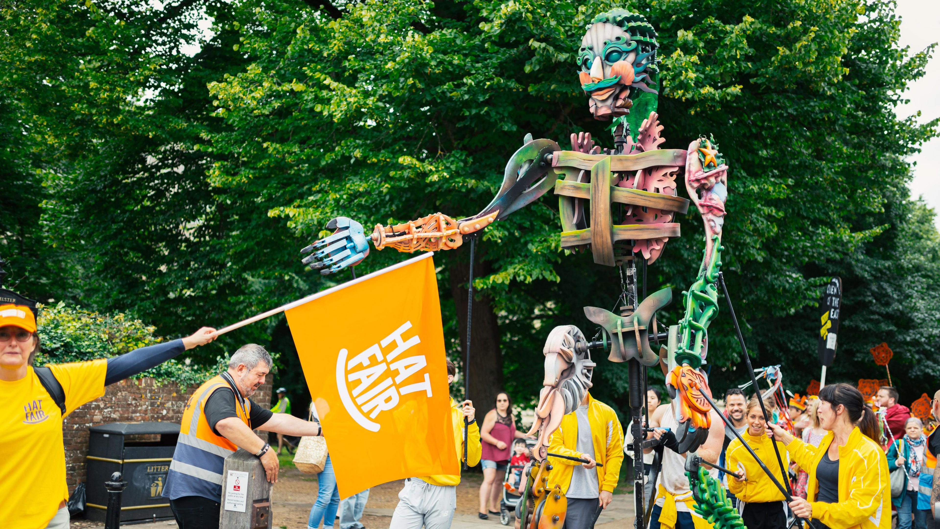 A puppet of a skeleton being controlled by multiple people in yellow tops walking through the streets. There is also a yellow flag being waved by one person and the flag has the words "hat fair" printed on it