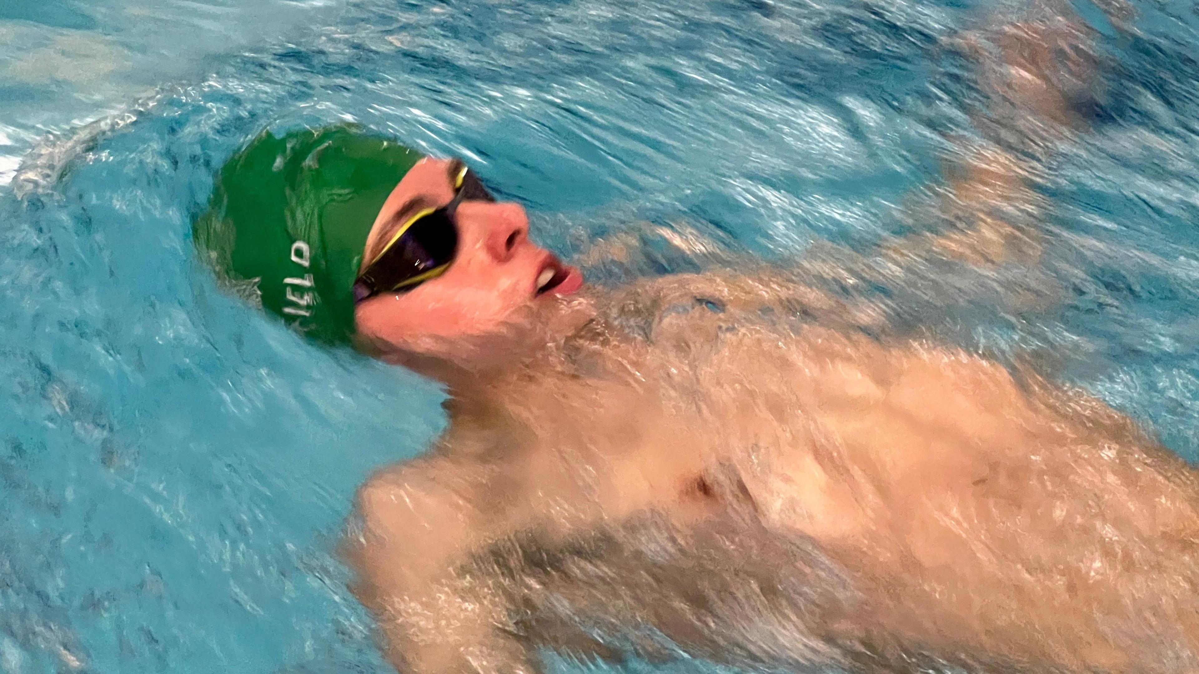 Filip is doing the backstroke in the pool. The photo is a close of Filip in the water looking up with his swim cap and goggles.