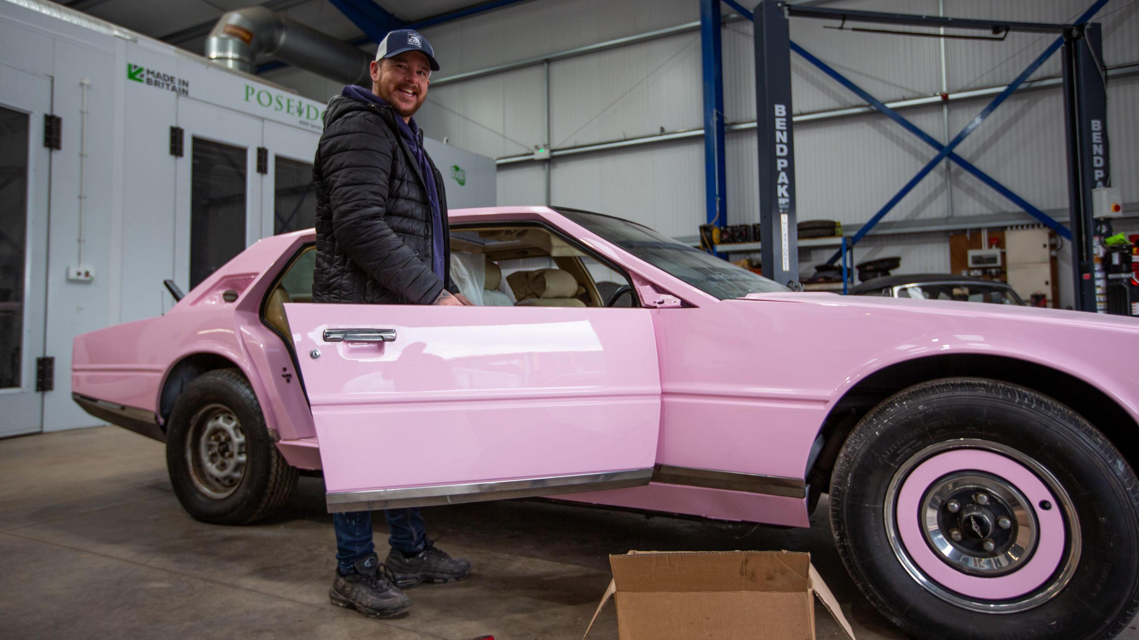 A man stands with the Aston Martin in the workshop after it was painted pink. The car and wheels are now pink and he is standing with the door open. He is wearing a blue and white cap and a black puffer jacket, with dark jeans and black training shoes.