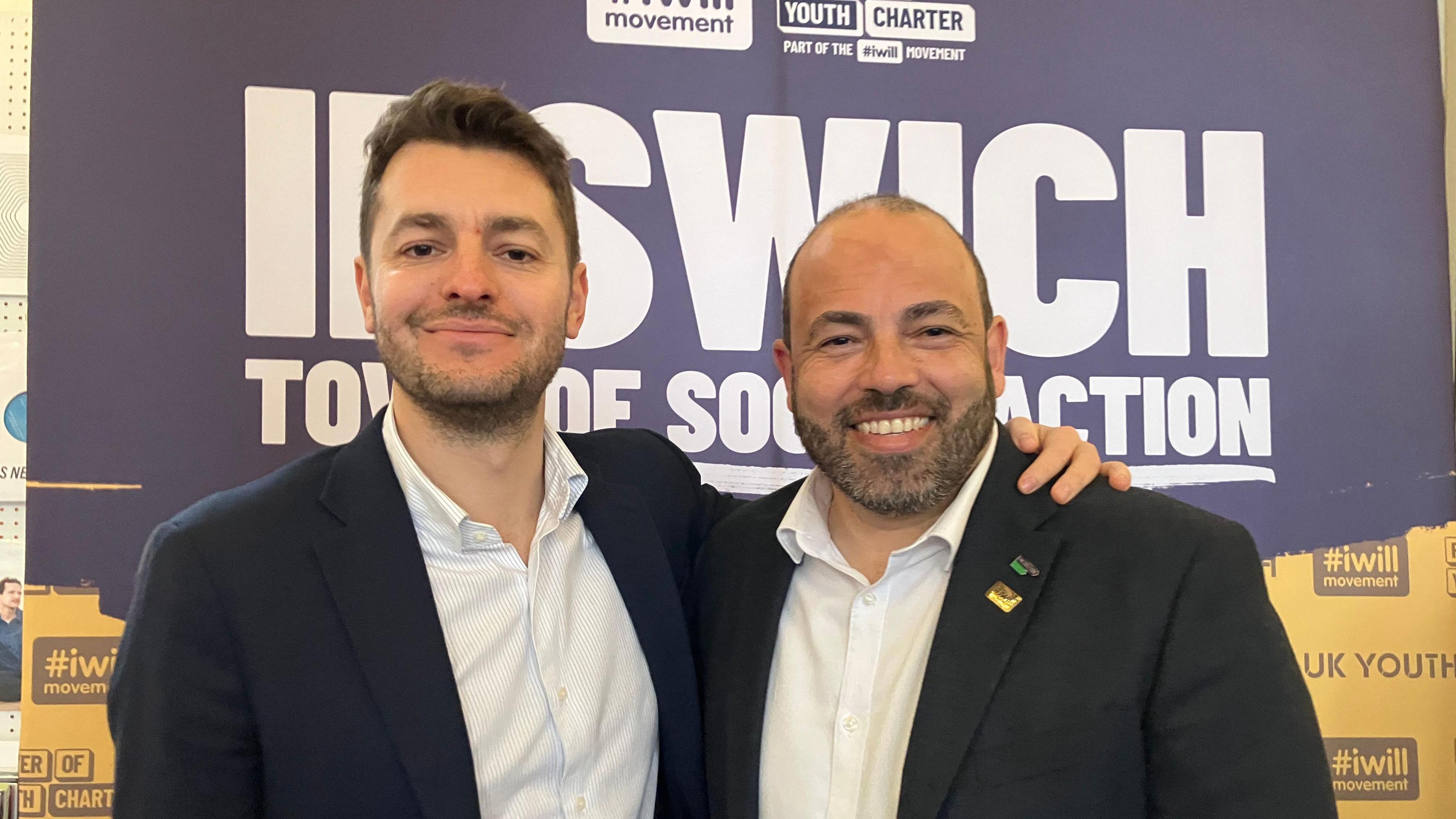 Two men in suits smile at the camera. They are in front of a banner which reads "Ipswich town of social action".