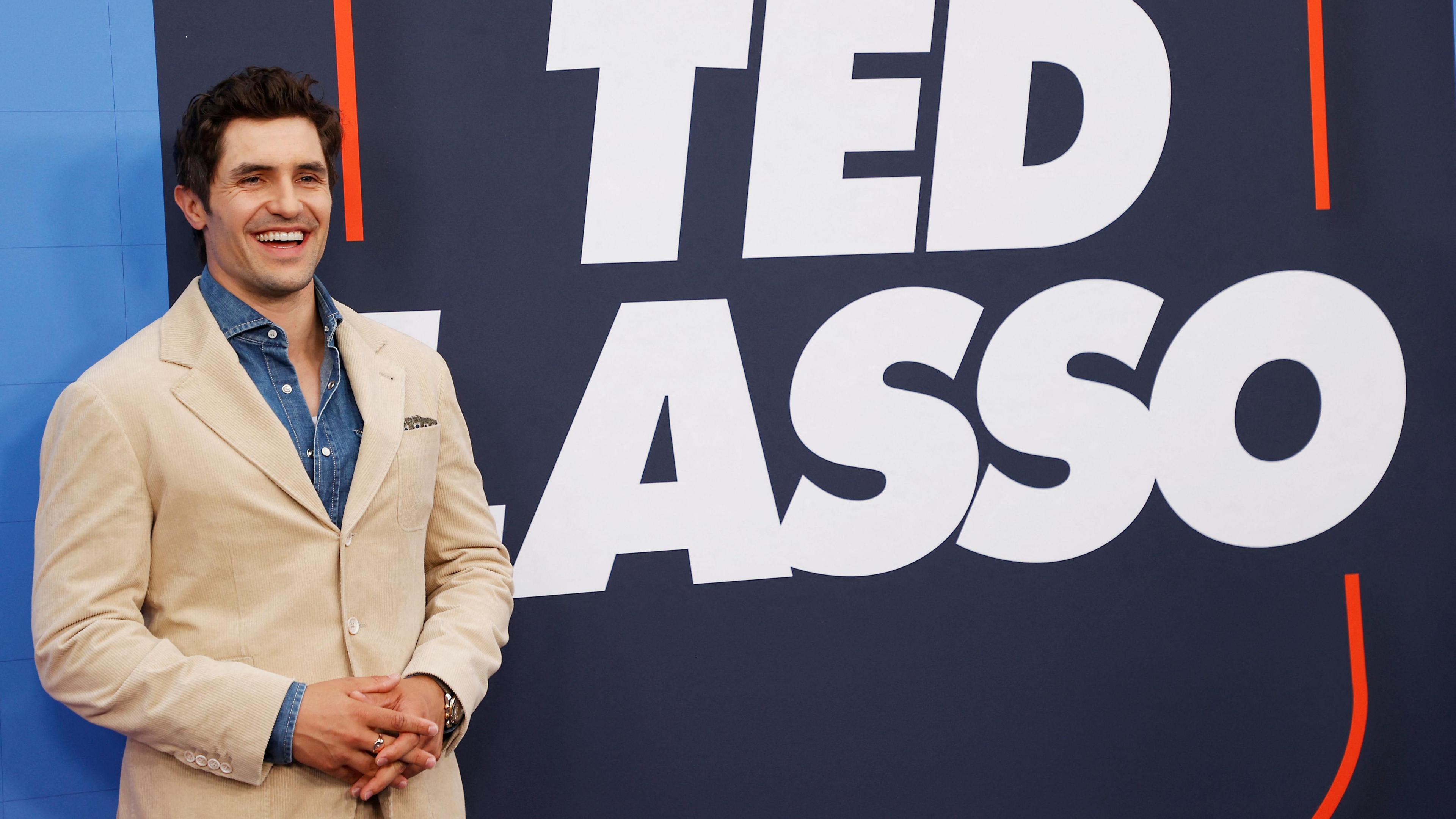 Phil Dunster, wearing a cream jacket and blue shirt, standing in front of a sign for TV programme Ted Lasso. He has dark hair and is smiling.