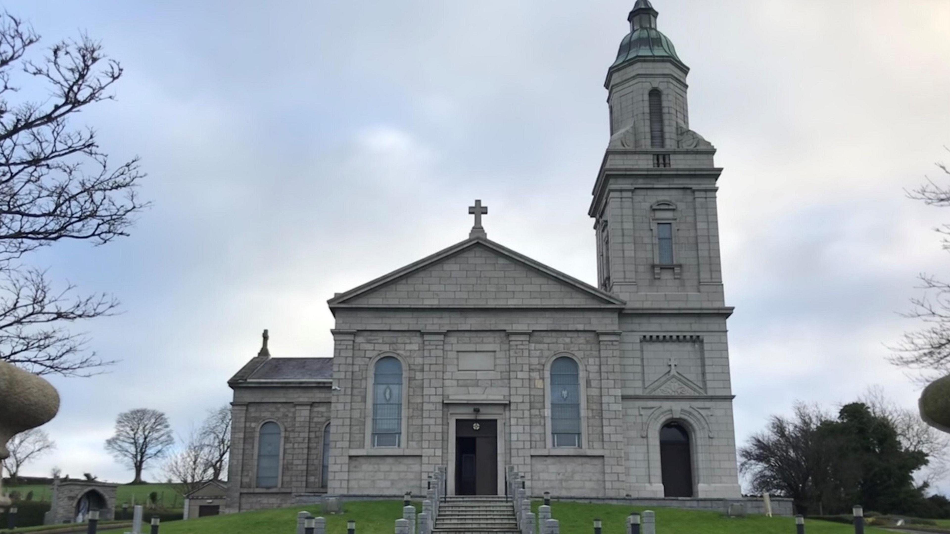 St John the Evangelist Church, Ballymaghery - a large grey building with a bell tower standing on a hill in front of steps and gardens.  A graveyard and trees are in the background.