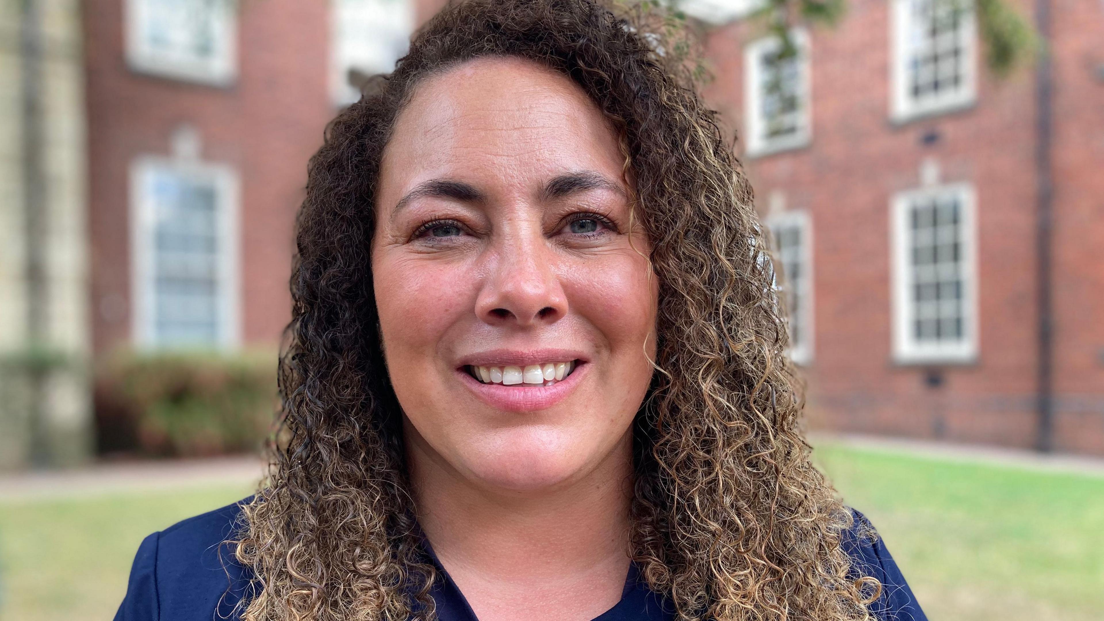 A head and shoulders photograph of Hayley Eccles. She has brown curly hair and is wearing a blue jacket. She is standing in a court yard.