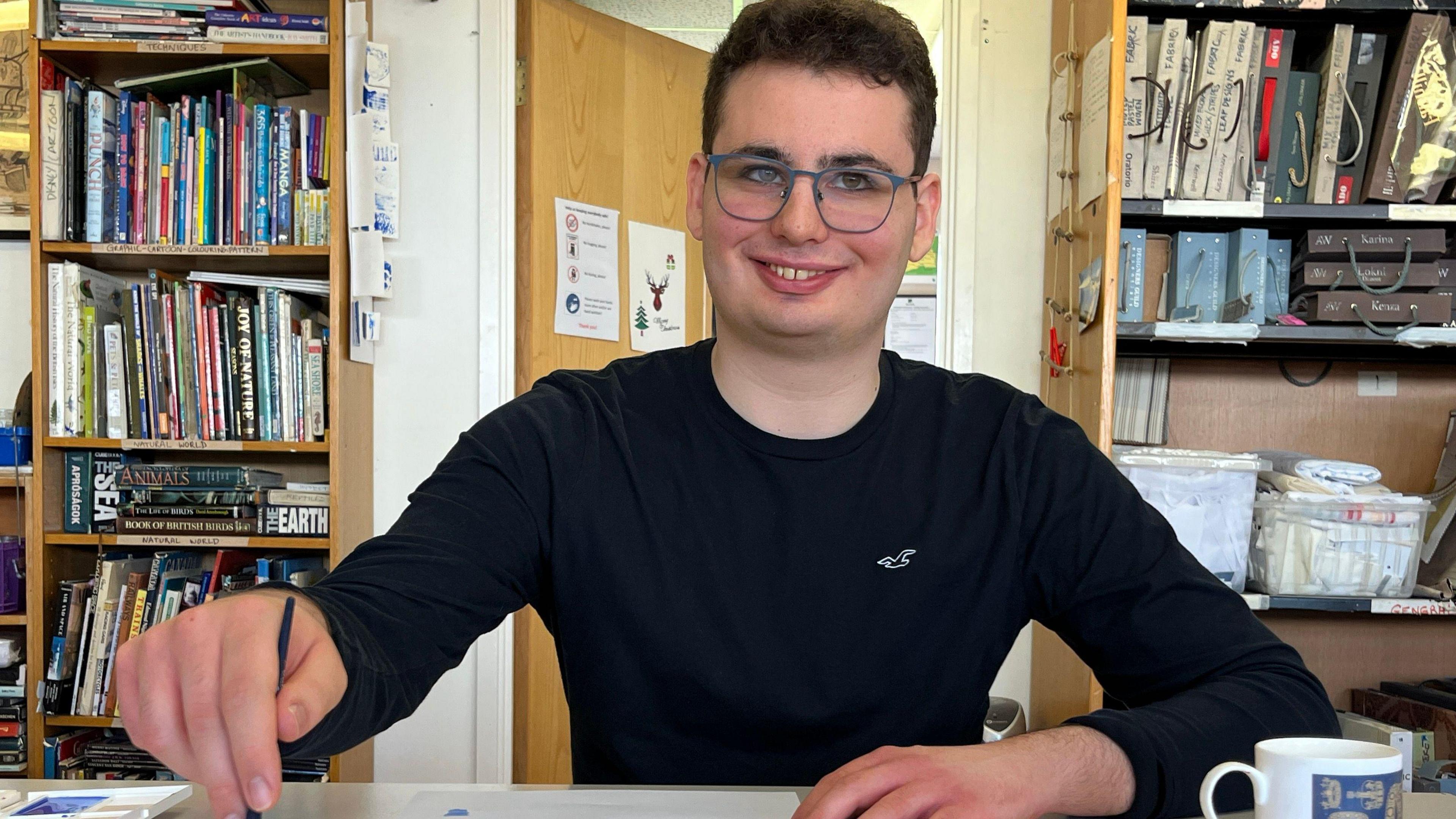 A young man dressed in black holds a paintbrush and smiles broadly at the camera