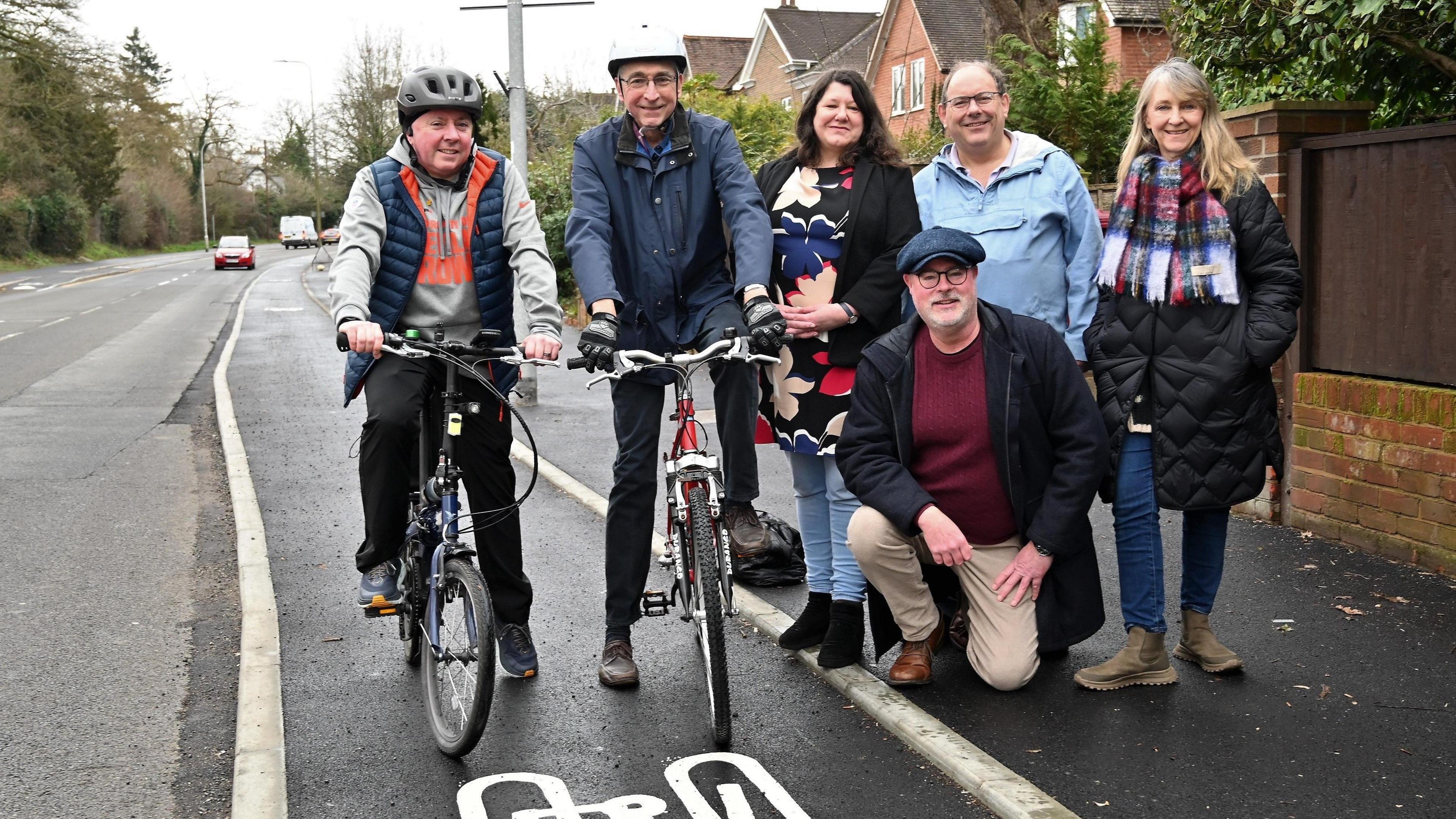 Two male cyclists on bikes posing in a cycle lane. They are standing with four other people, who are on the pavement.