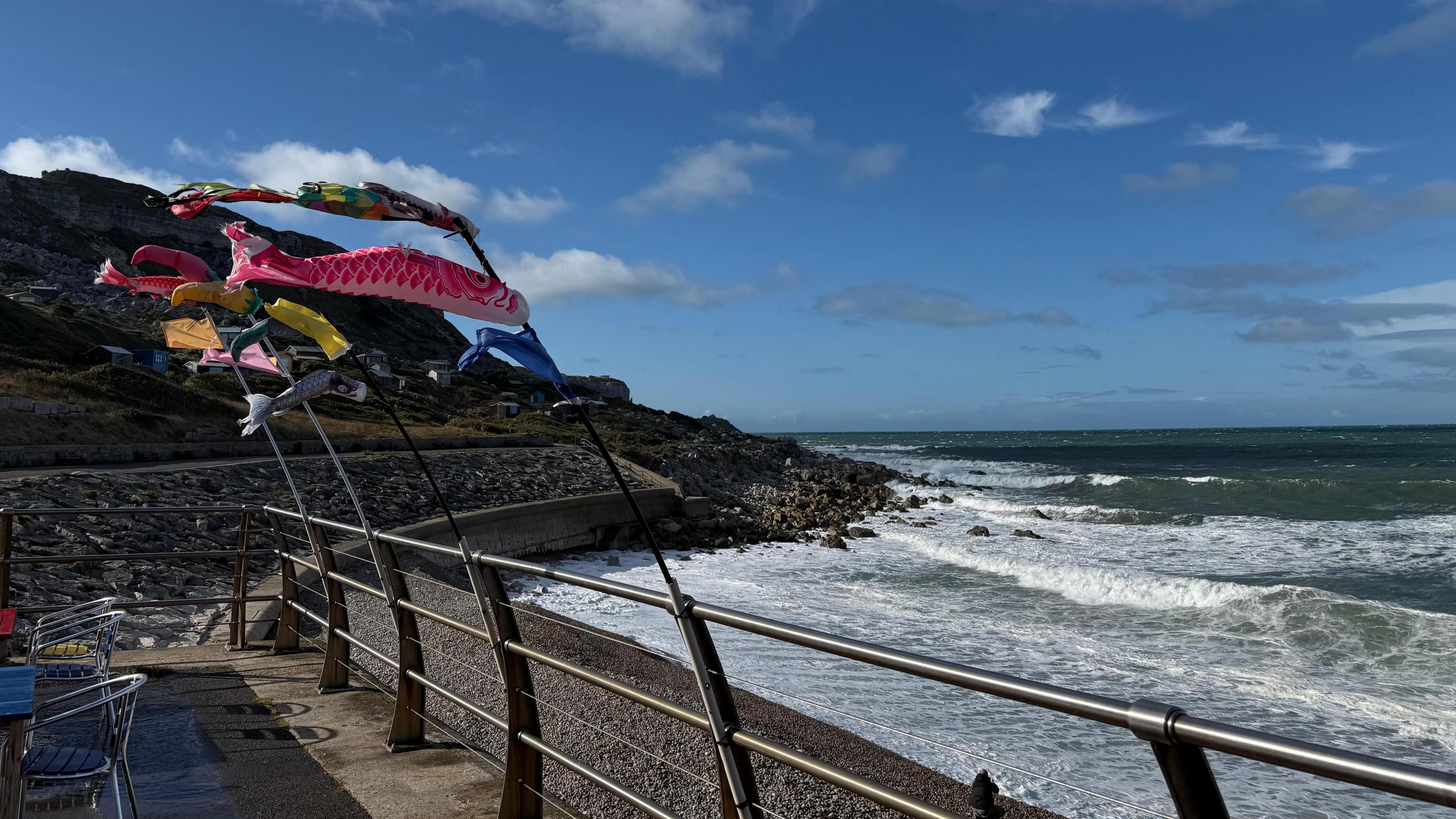Waves crash against a sea wall and rocks as a stiff breeze holds fish kites tied to poles out horizontally. The fish kites are multicoloured and fly just above some café chairs sitting on a terrace. The sky is mostly clear bar a few sparse fluffy white clouds and the sun is shining.