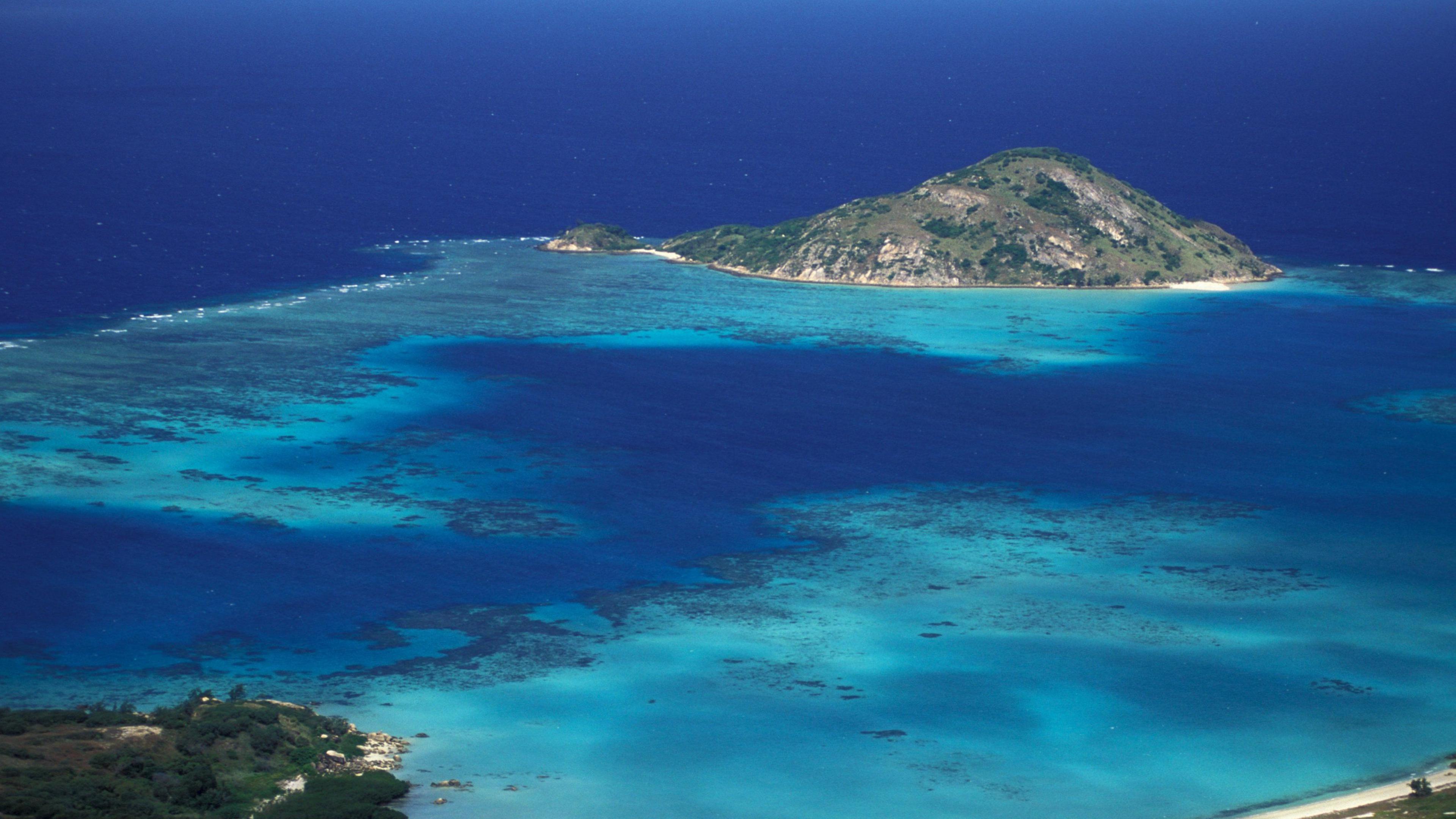 An aerial shot of Lizard Island - a small island on Australia's Great Barrier Reef - showing the calm blue shallow waters of the reef. The island is dominated by a peak.