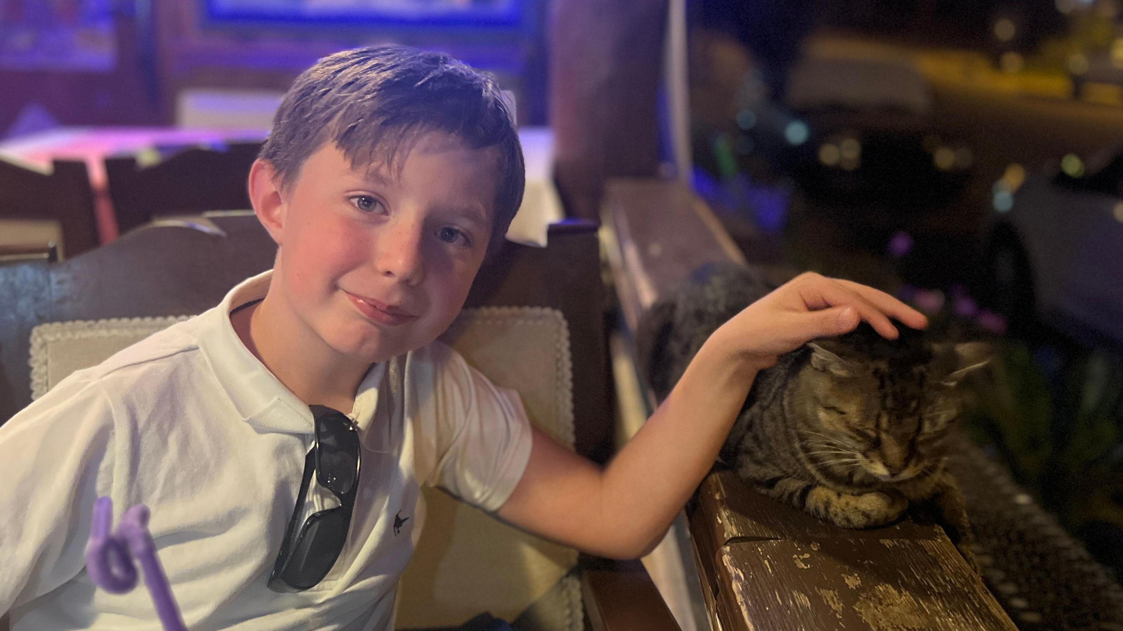 A young, smiling boy sits in a chair with sunglasses tucked into his white T-shirt. He has a hand on a tabby cat that is resting on a brown ledge. A purple straw is in the foreground. 