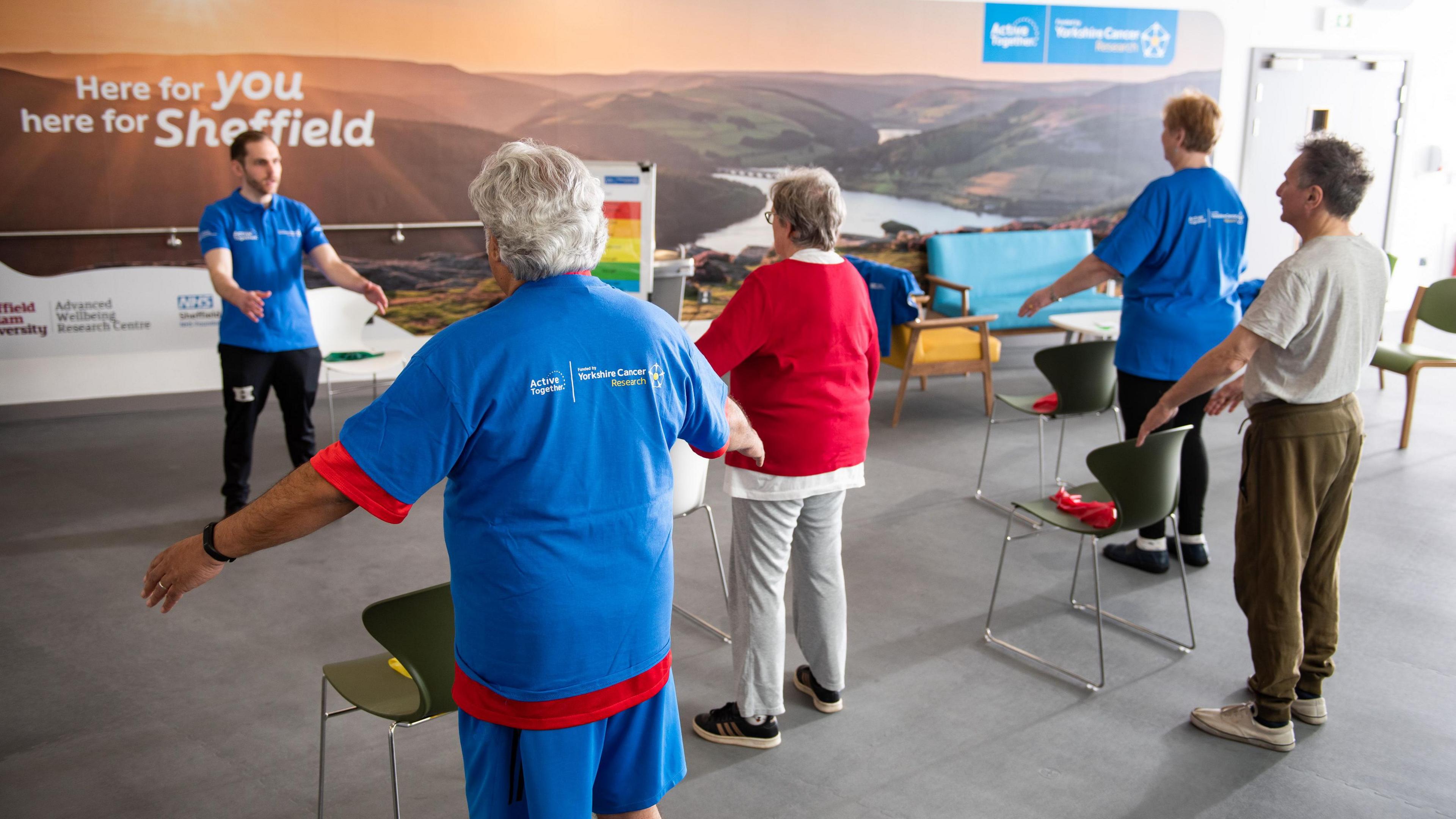 A picture of five people stood in a room with their arms outstretched doing exercise. 