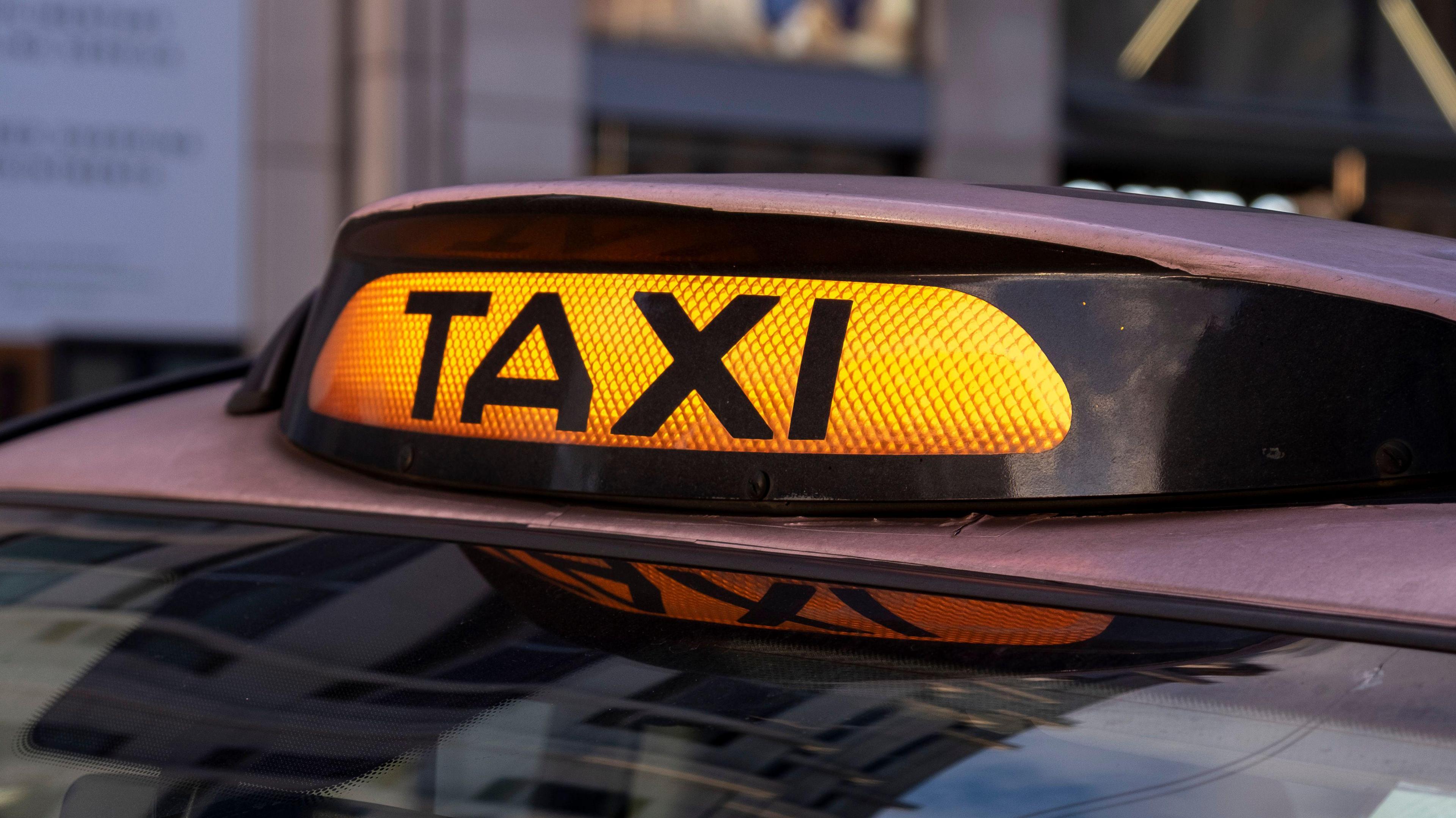 An orange illuminated "Taxi" sign on top of a black cab