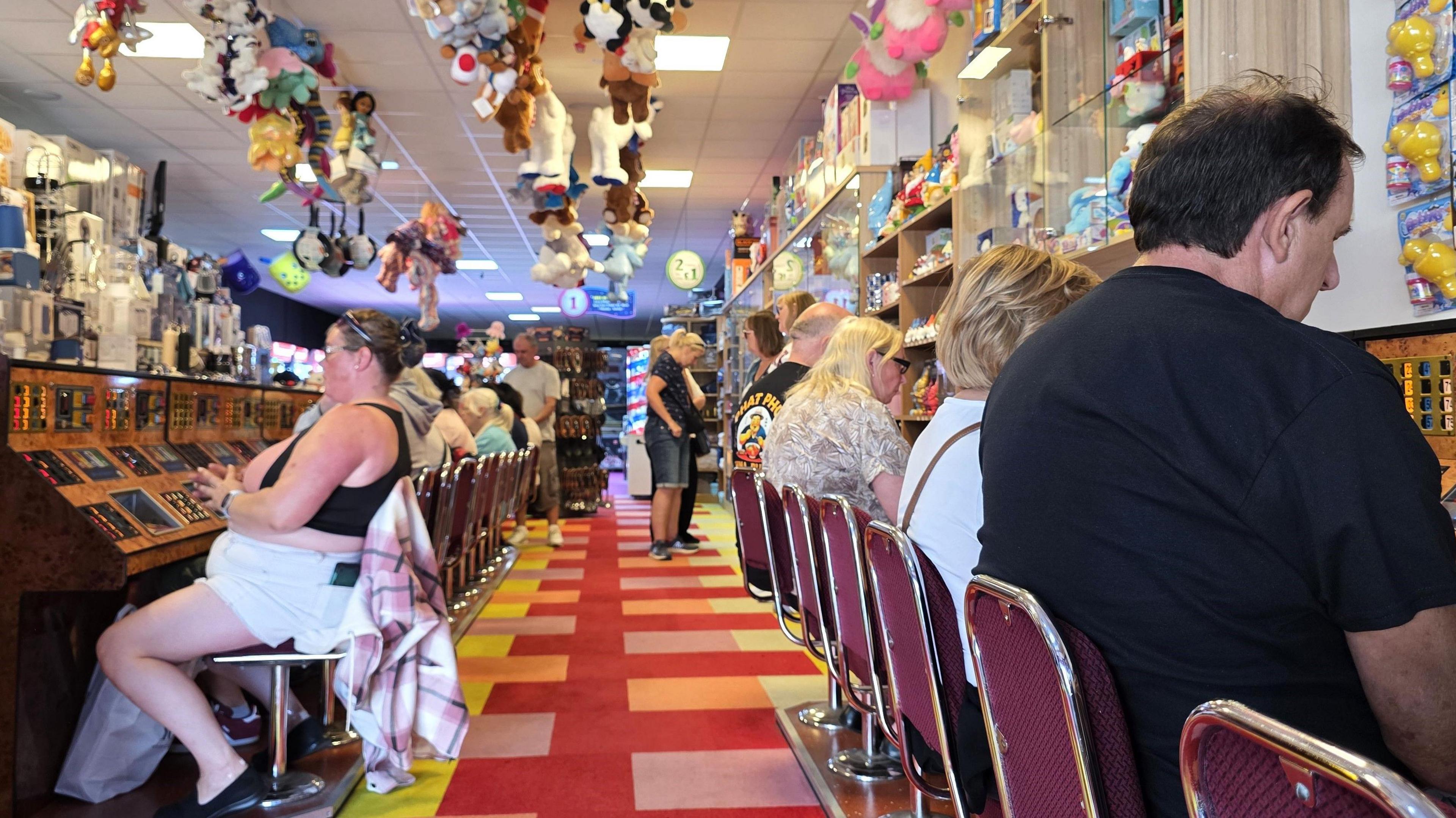 We can see several people sat down and playing bingo. Above them and around them are a wide range of prizes, including cuddly toys.