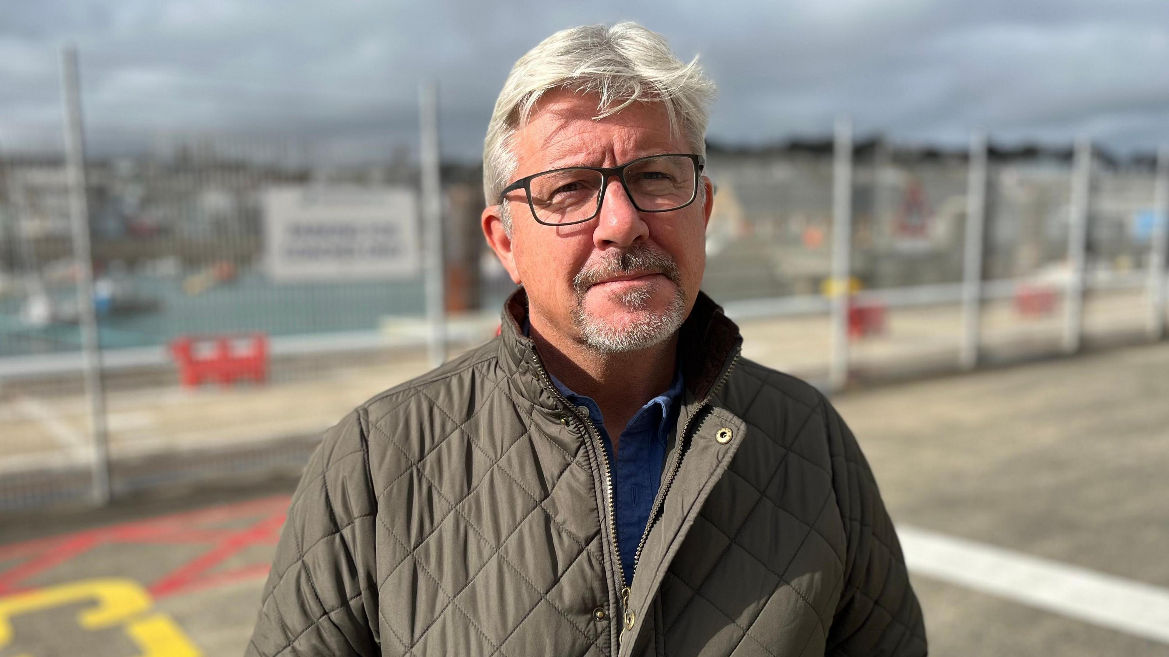 A man wearing a green jacket looking towards the camera. He has a grey beard and grey hair. 