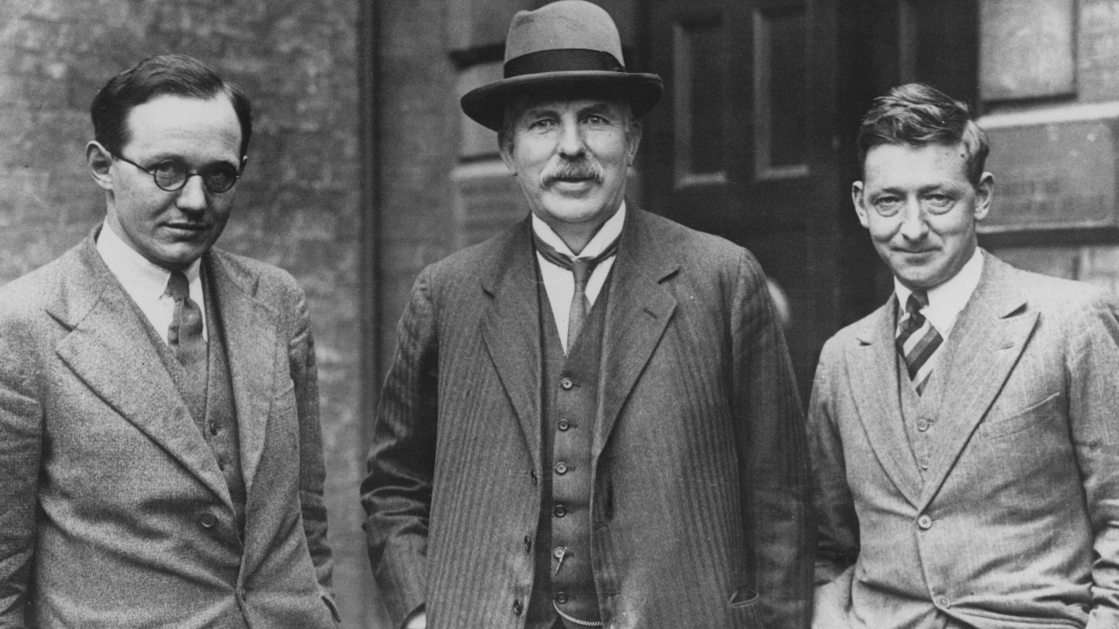 Black and white photo showing Ernest Walton, Sir Ernest Rutherford and James Cockcroft in suits and ties, standing in front of a door