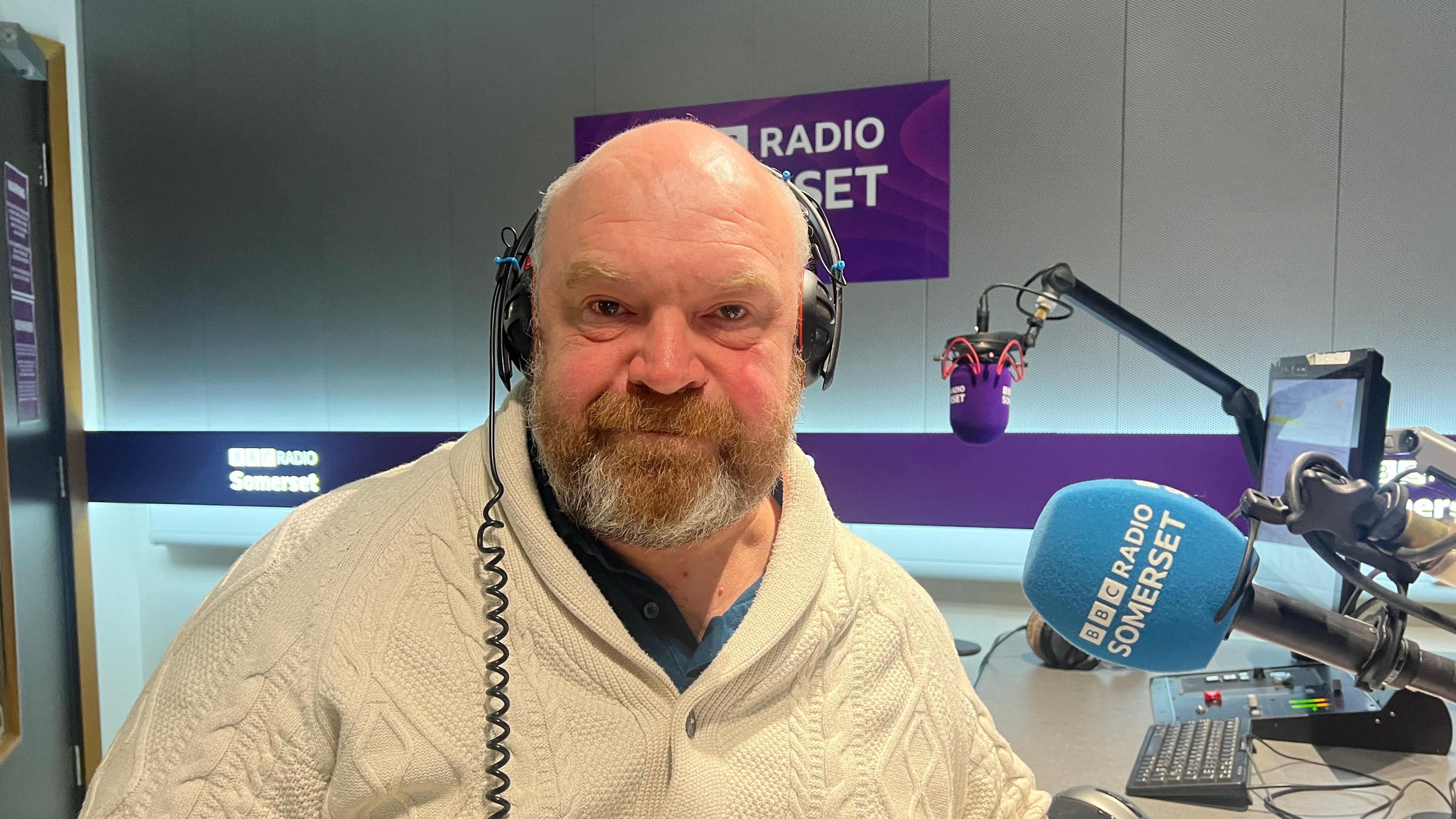 A man wearing a woolly cream knitted jumper and black headphones is facing the camera. He is in a radio studio with a blue BBC Radio Somerset microphone next to him.