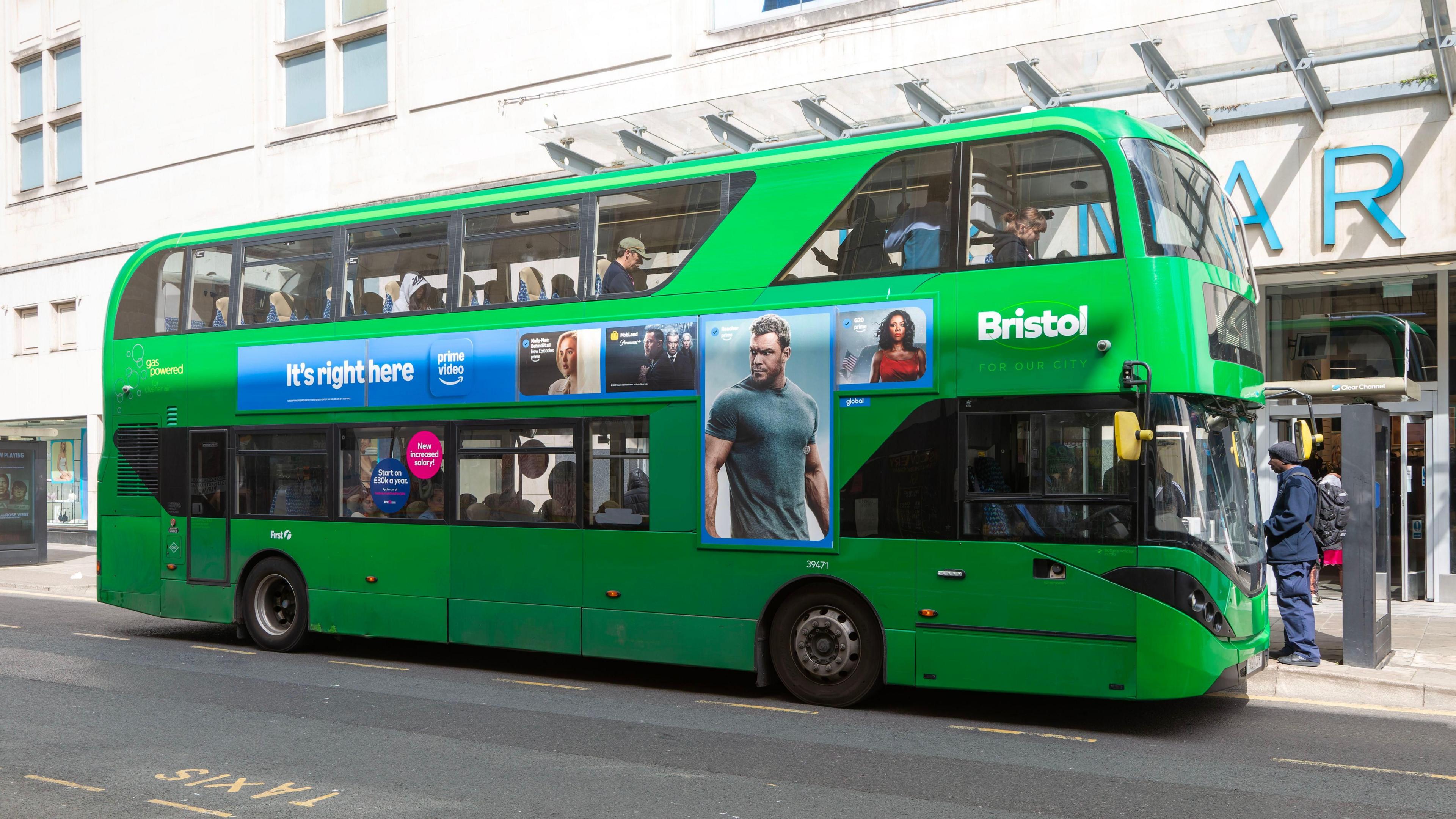 A green double decker bus at a bus stop outside Primark in Bristol city centre. There is a person standing at the stop waiting to board.