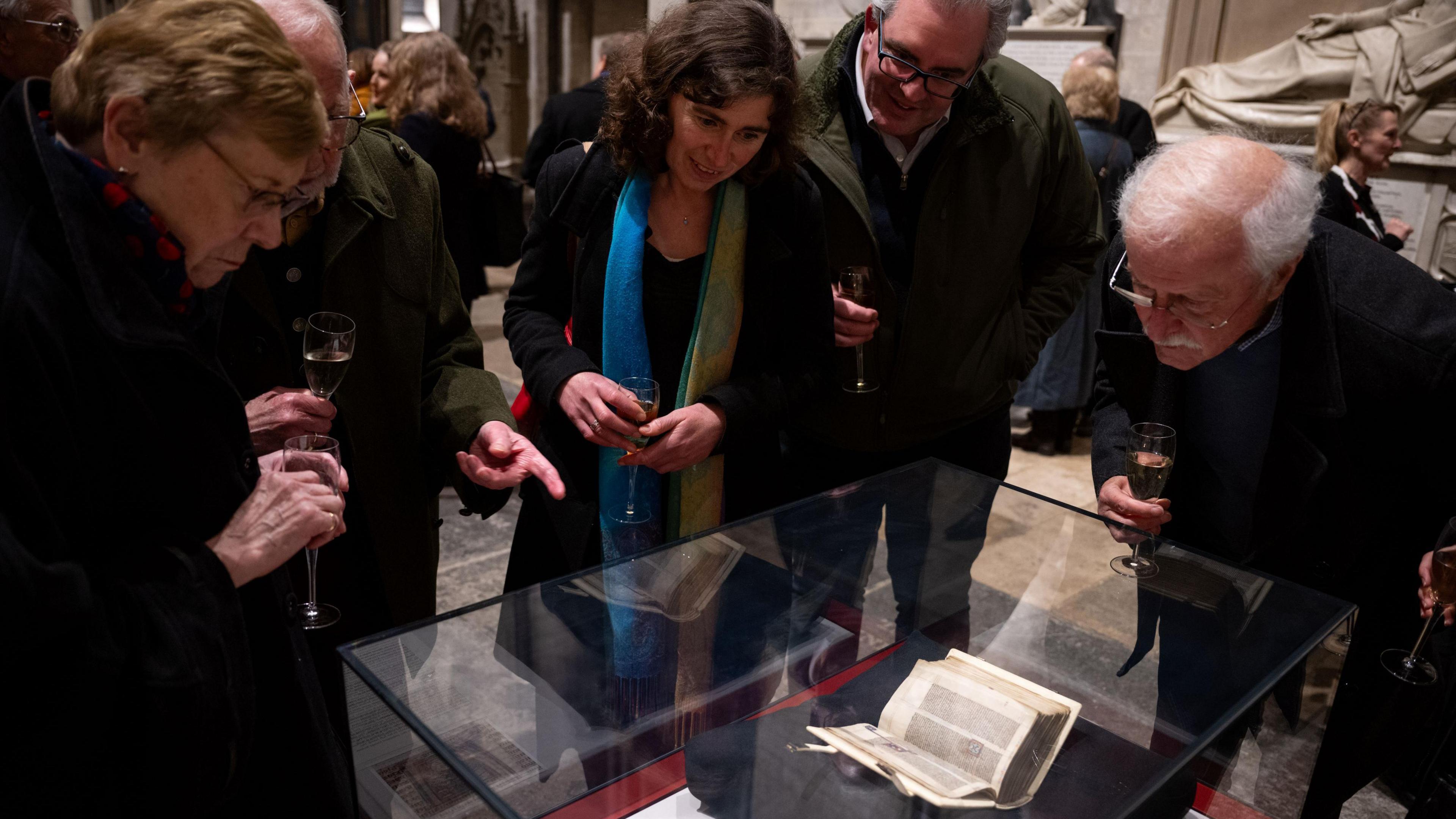 Several people holding glasses of bubbly peer at a glass case with a small open book inside. 