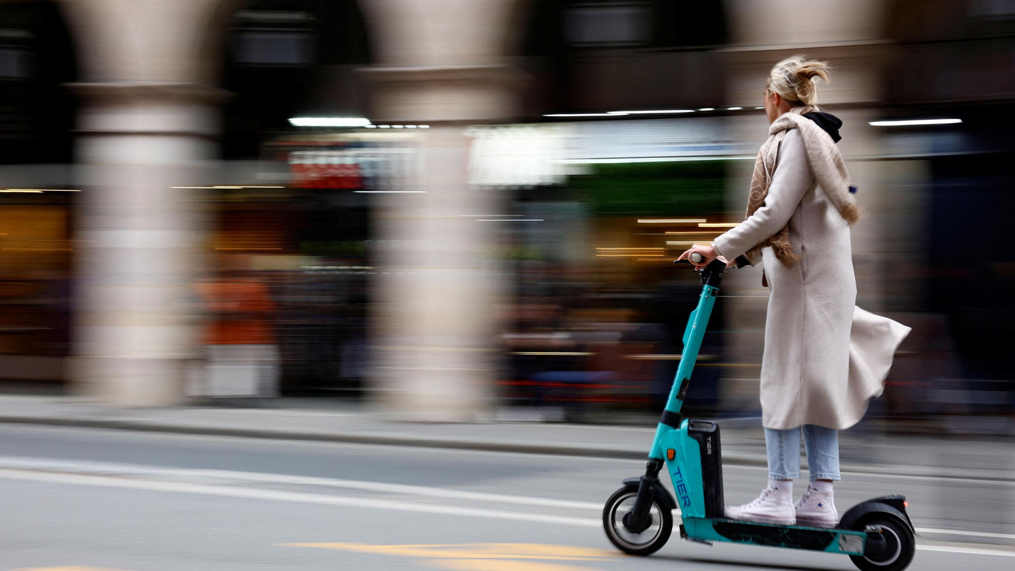 Woman in cream coat riding teal Tier scooter on road.