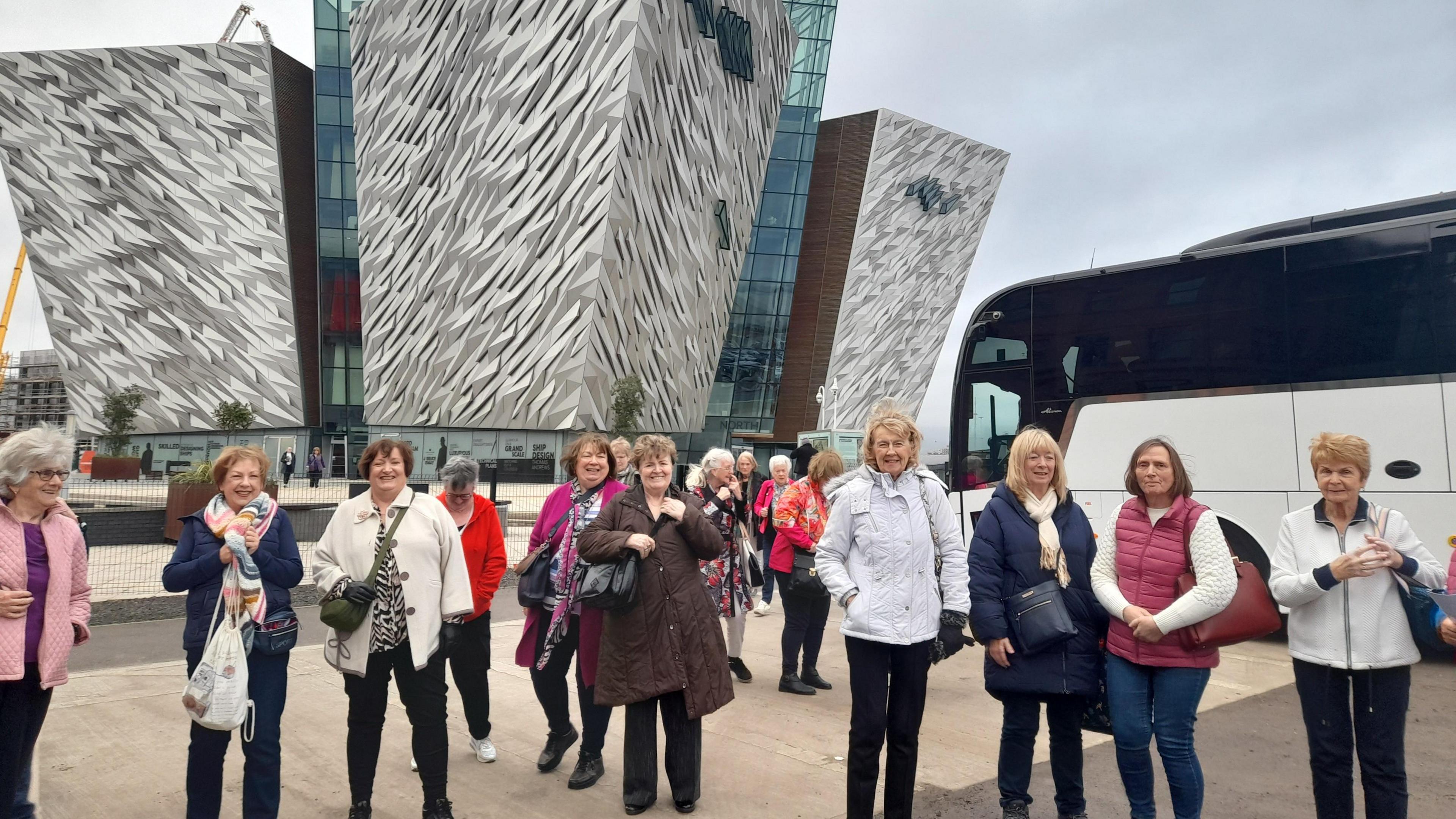 A group of women stand in front of the Titanic Belfast building. There are about a dozen women wearing winter clothing. Behind them, beside Titanic Belfast, is a white coach.