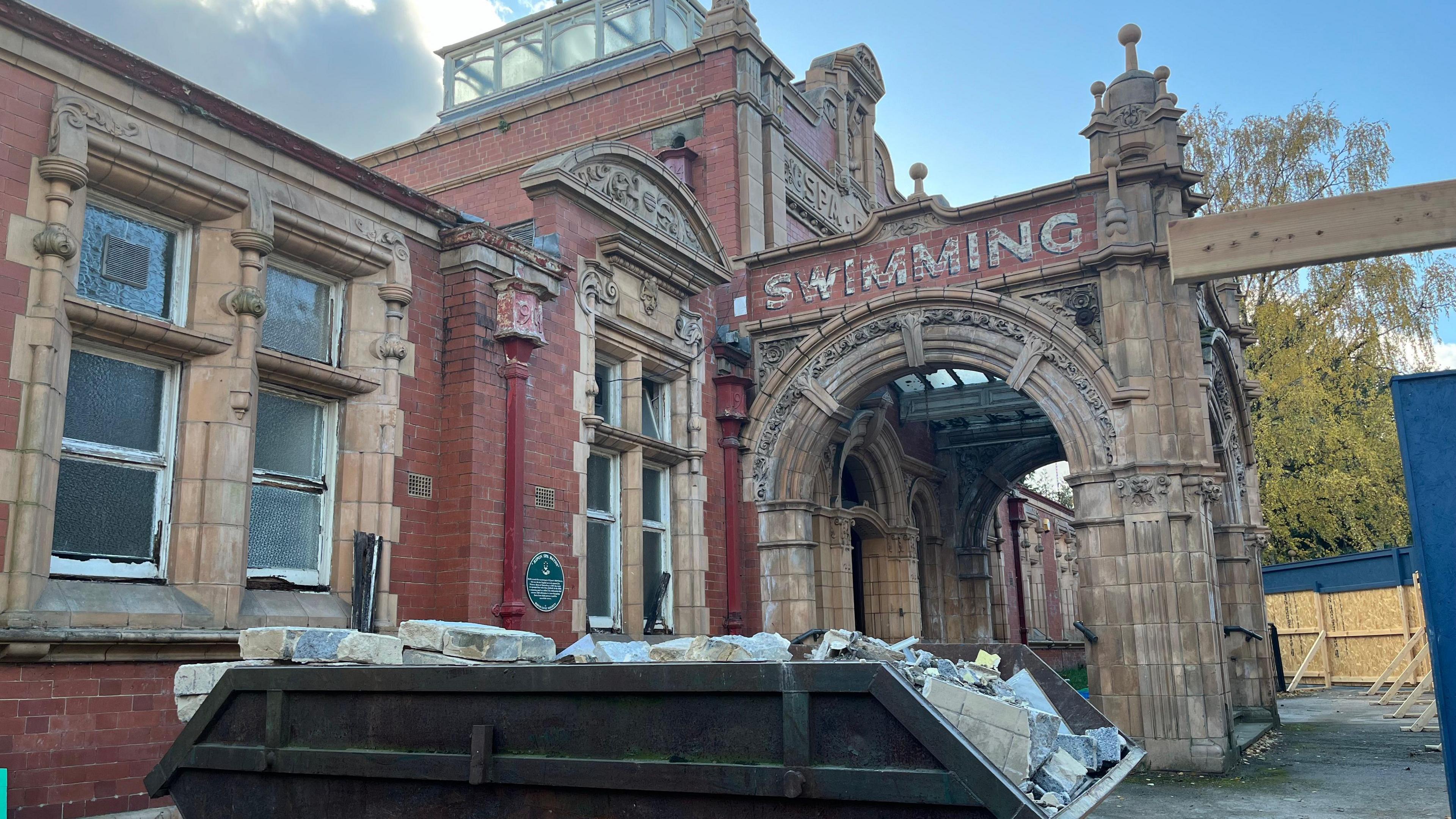 A brick building with archways and the word "Swimming" written on the side.  A full skip is in the foreground.