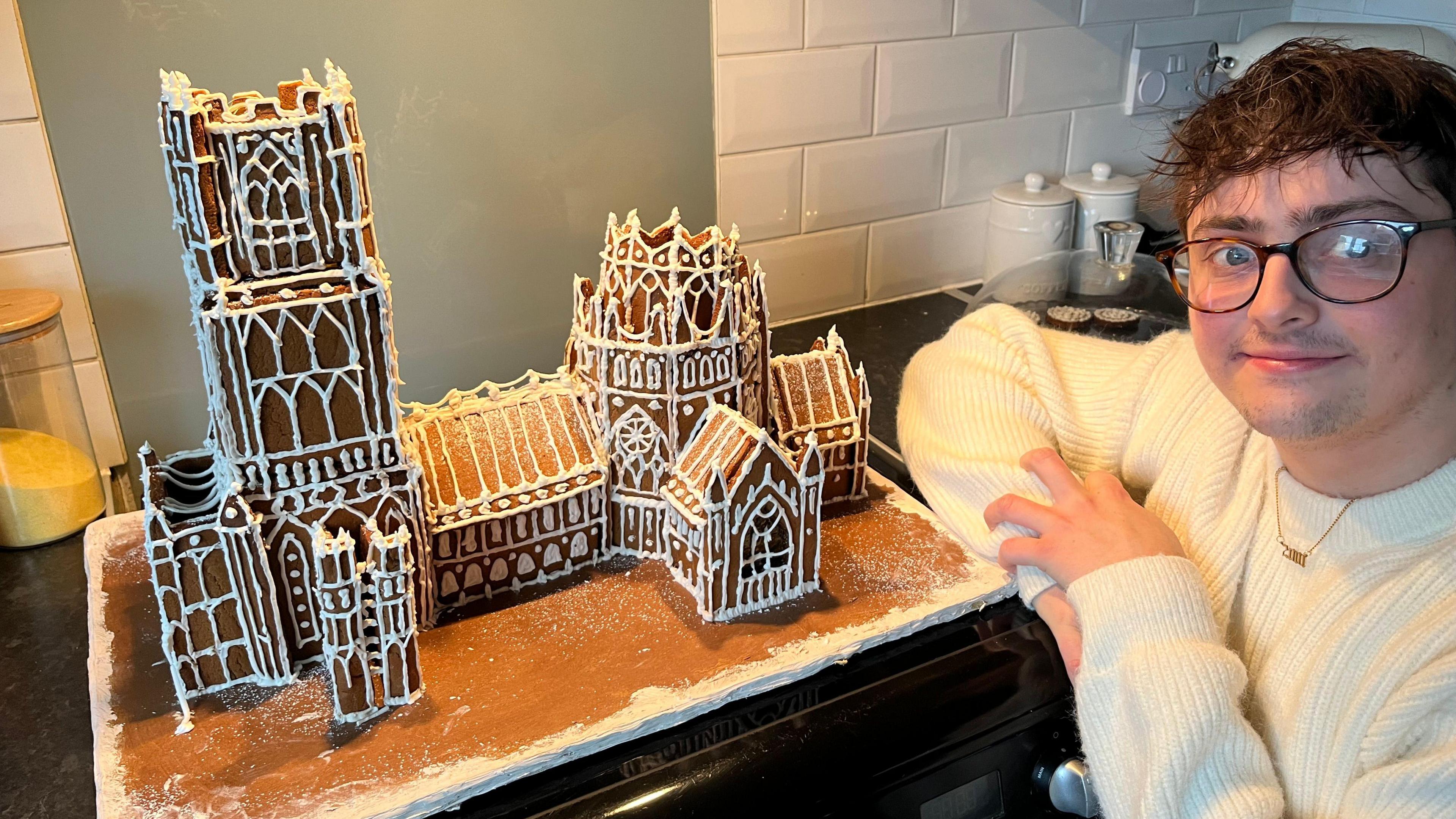 Owen Angier in a white woollen jumper, he is to the right of a model of Ely Cathedral made from ginger bread, with white Royal icing piped around windows and doors. Owen has dark hair across his forehead, and wears glasses with black frames. A white tiled kitchen wall is behind him and a grey splash back guard is behind a cooker hob.