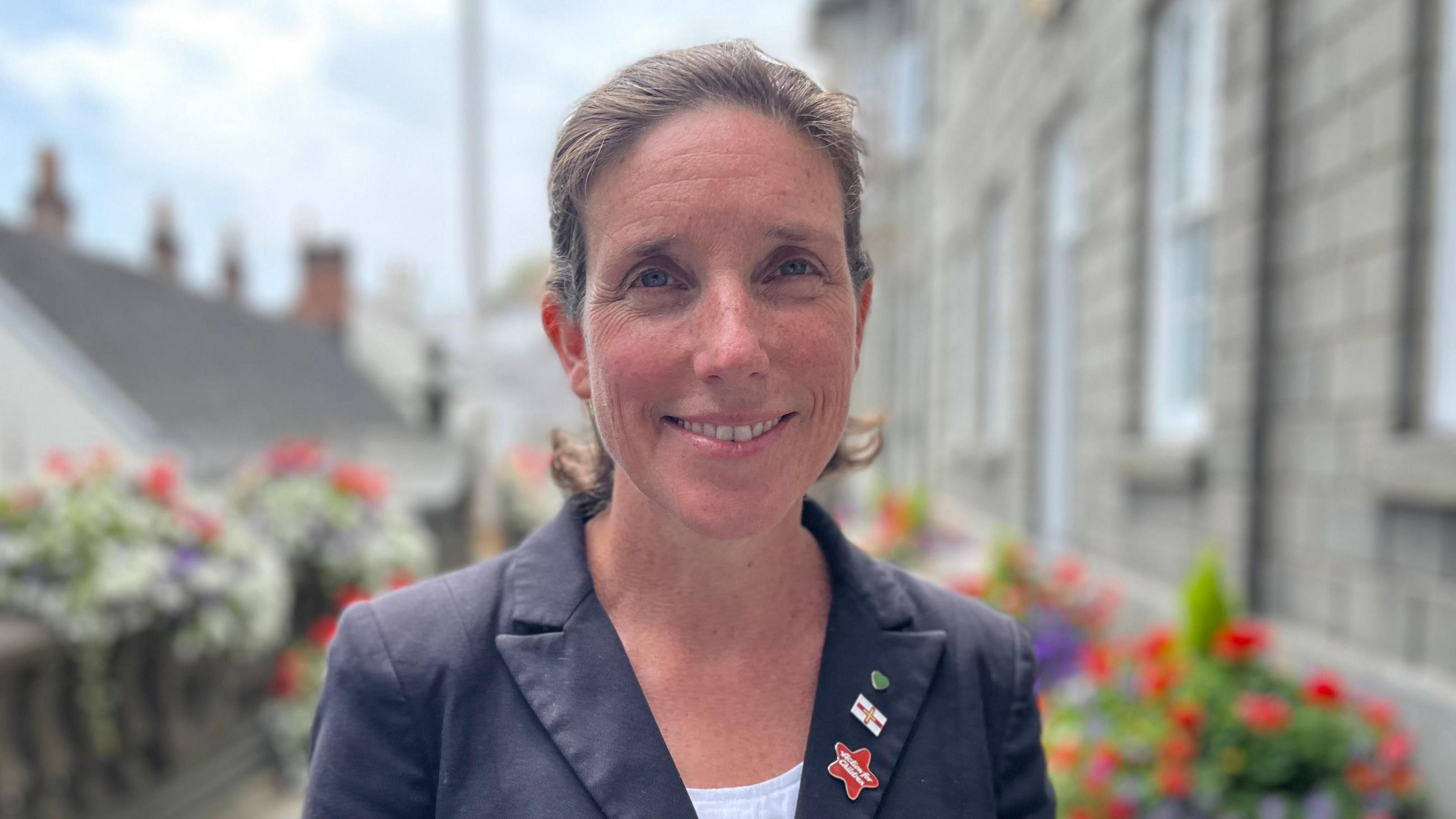 Deputy Lindsay de Sausmarez, a woman with brown hair wearing a blue jacket and three pun badges on the lapel. She is smiling towards the camera.