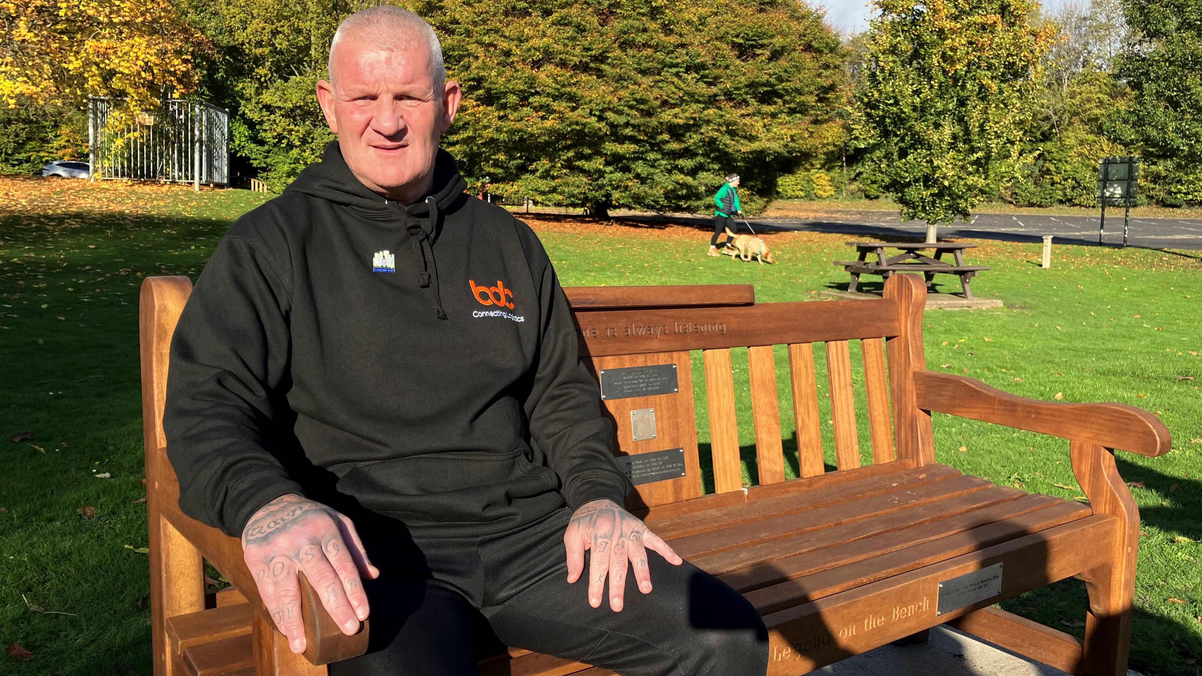 Man with dark sports suit sat on a bench in grass. There are trees behind him.