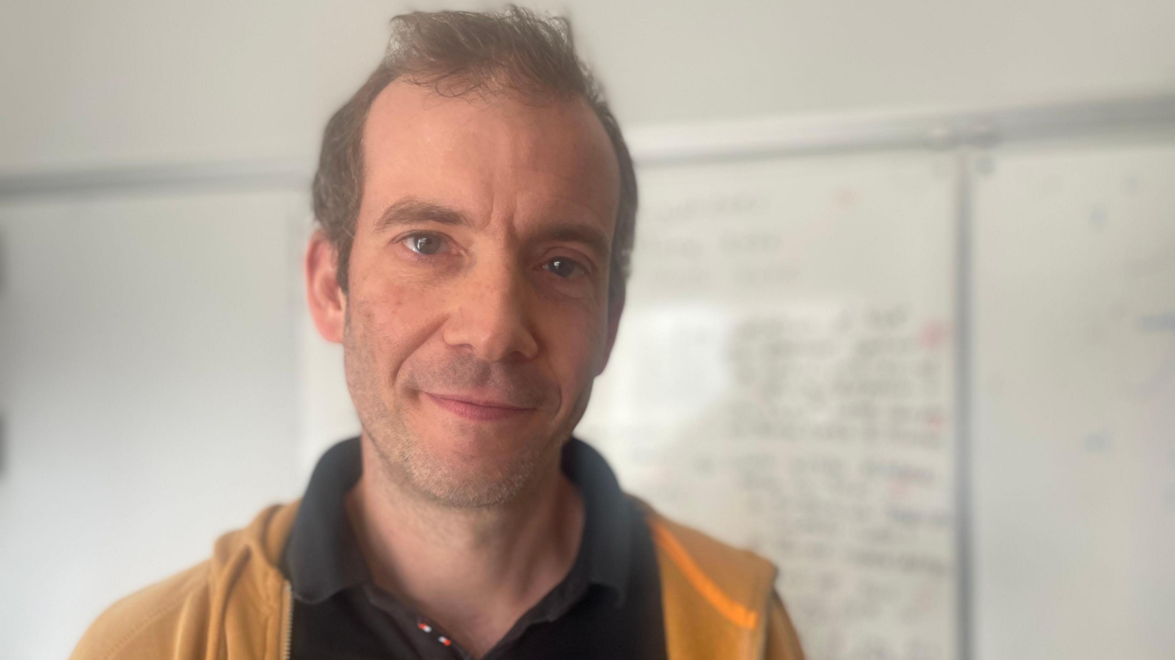 A man with short brown hair wearing a black polo shirt and yellow hoodie standing in front of a white board with writing on it.