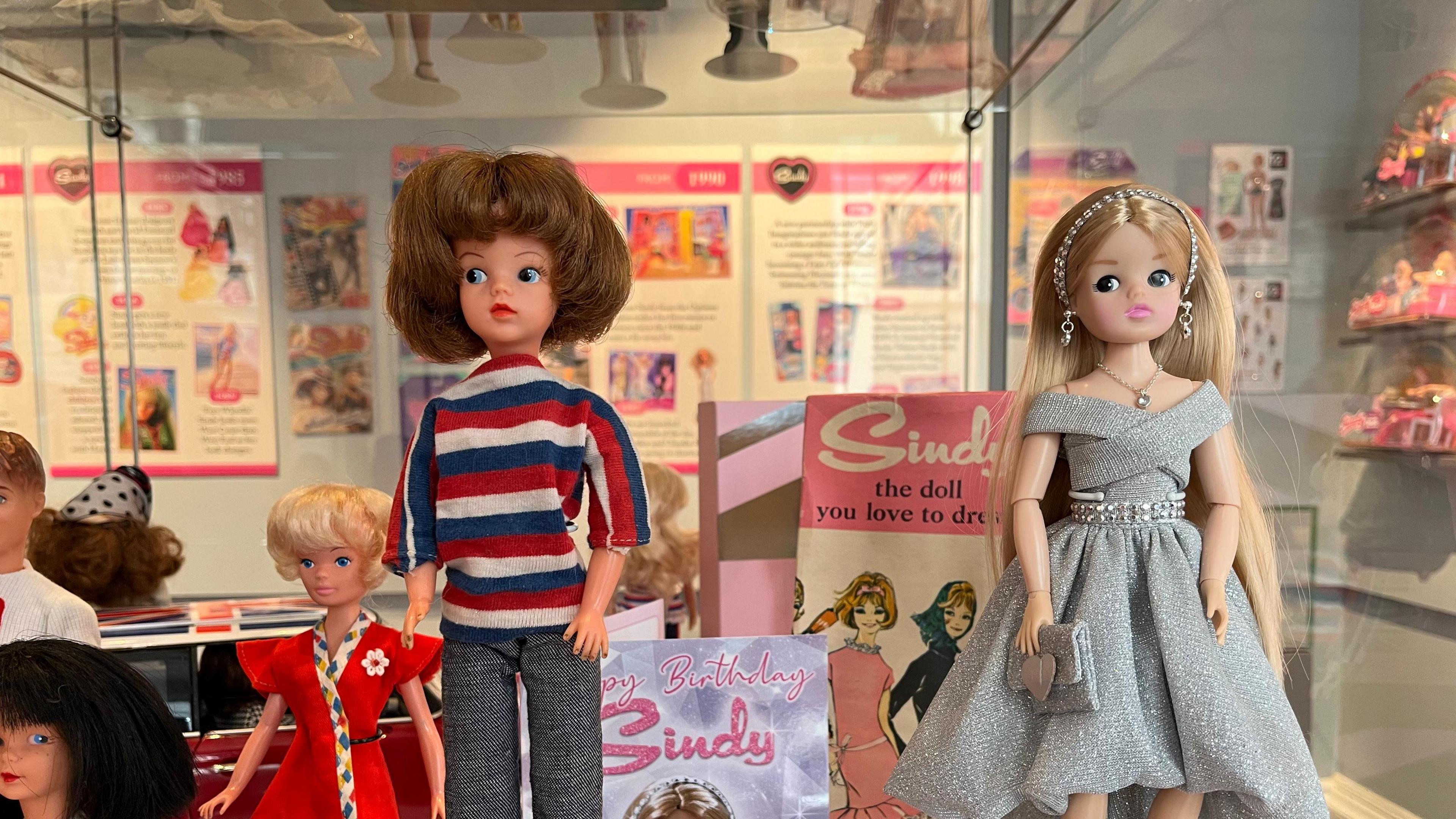 Two Sindy dolls in an exhibition case. The doll on the left is wearing a striped top and grey trousers and has brown bobbed hair. The doll on the right is wearing a silvery dress, off the shoulder and has blonde hair. Signs in the case say Sindy the doll you love to dress. Chippenham Museum, 2024