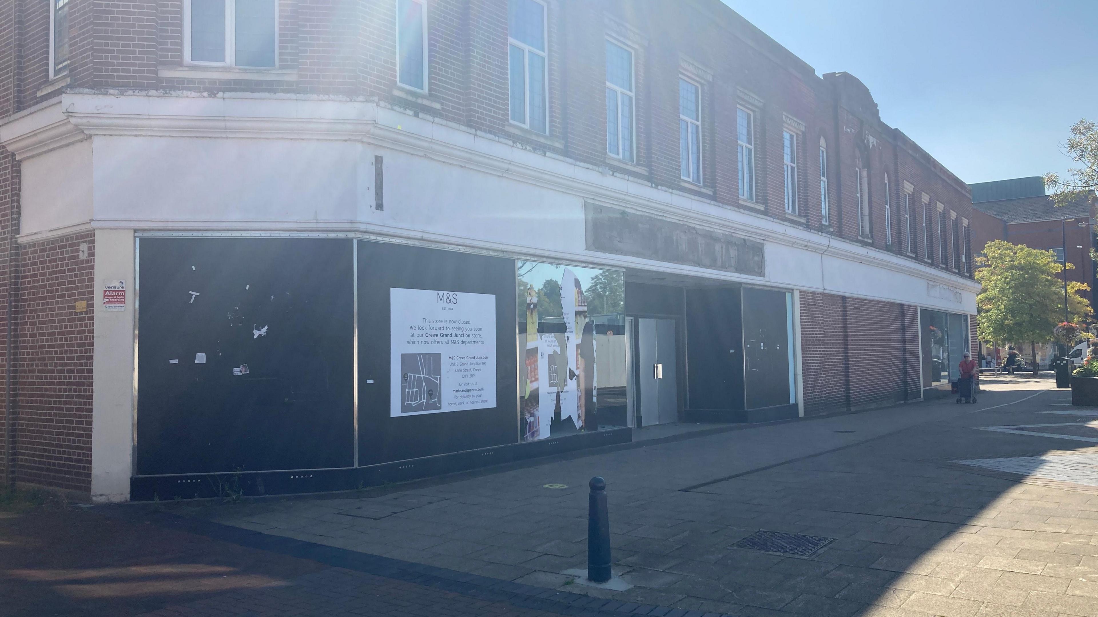 The unused M&S store in Crewe town centre. The outside has boarded over windows in front of a paved area with a bollard in it. A sign on the outside has the M&S logo on it and text underneath. Sunlight is reflecting off windows above the facade.