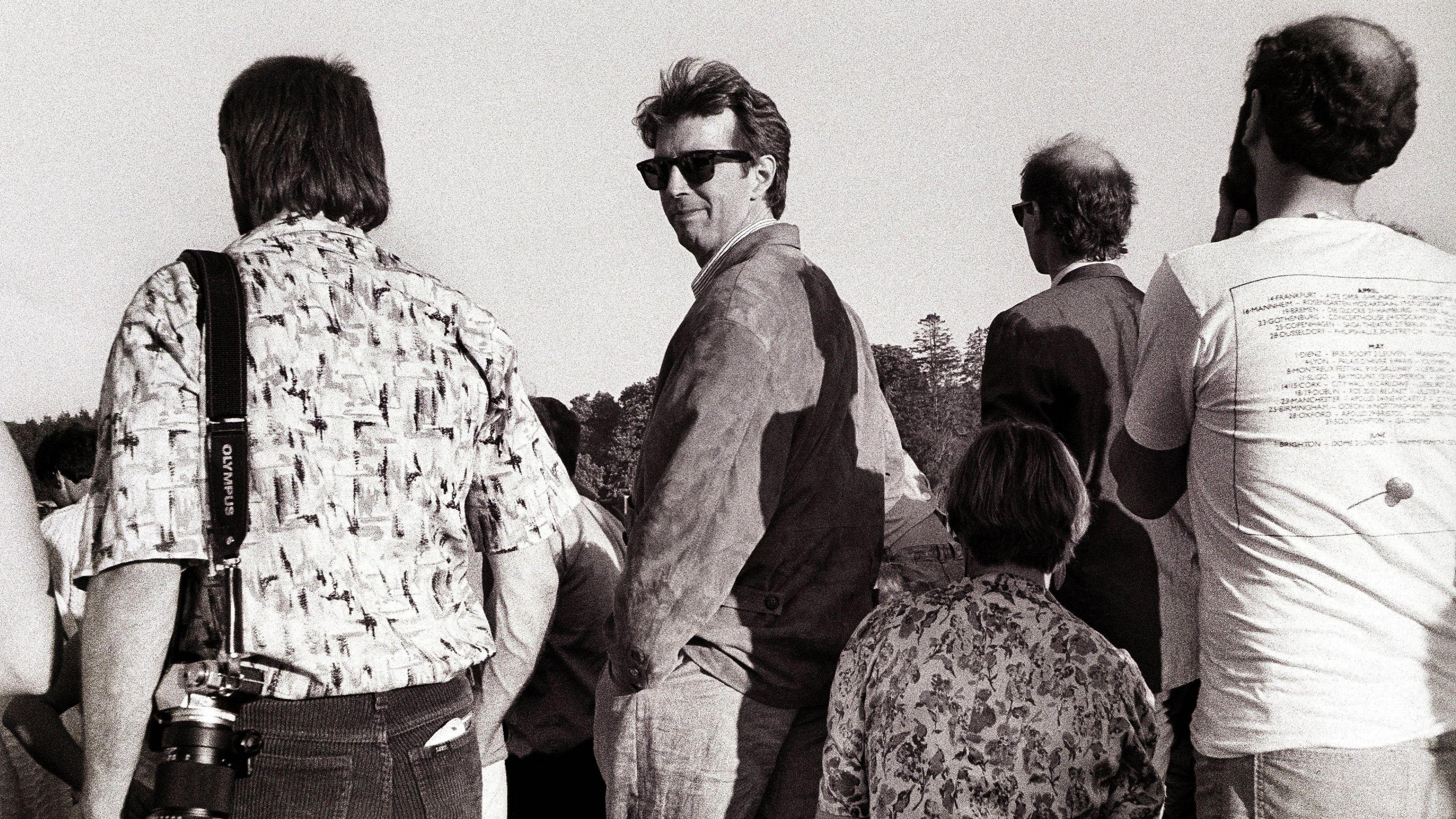 A black and white image of Eric Clapton turned around in a crowd wearing sunglasses as others are facing the opposite direction. Eric has short dark hair and is wearing light trousers and a light jacket. 