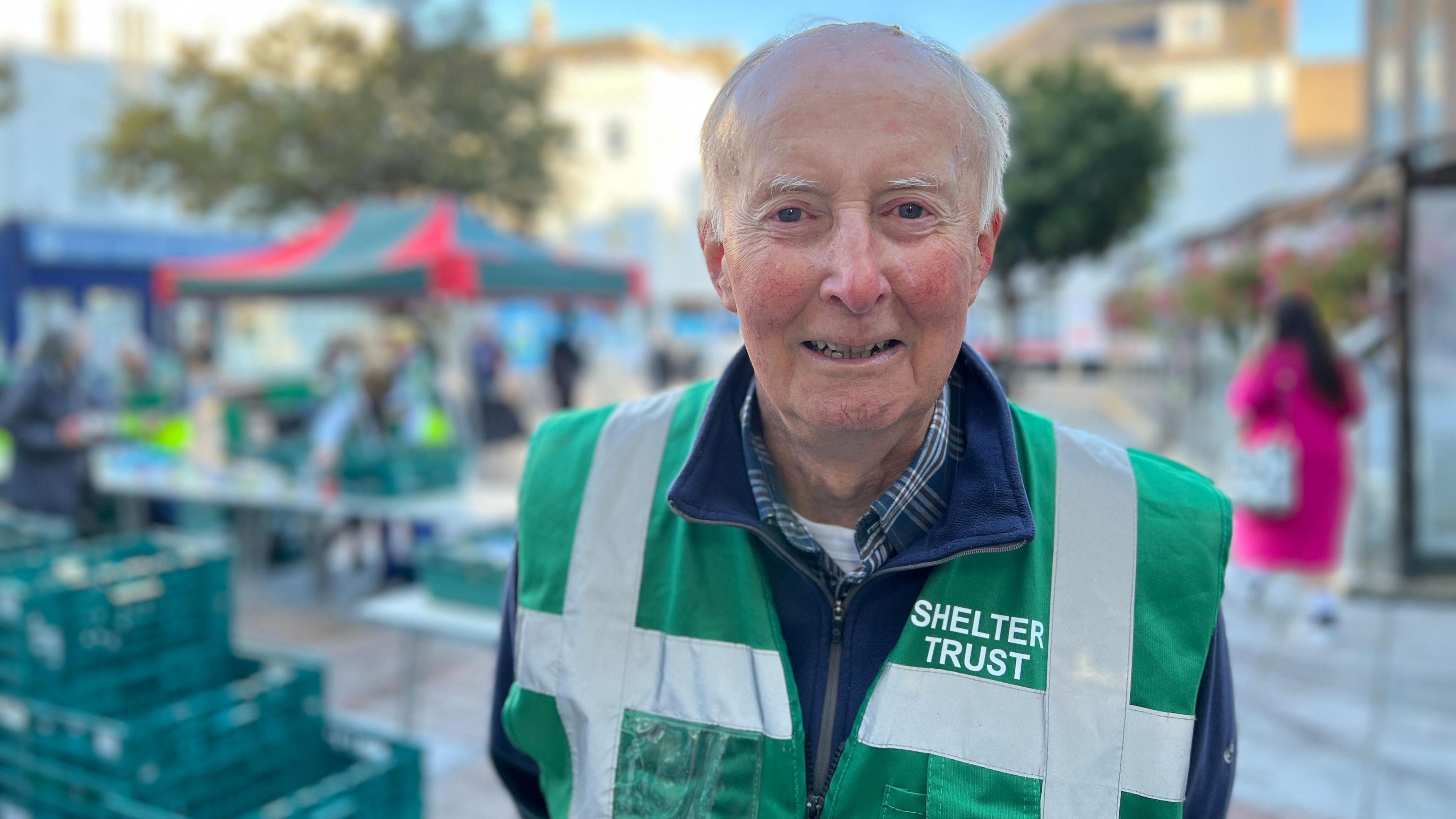 Richard smiles at the camera and is wearing a blue jacket with a shirt underneath it. He has a green bib on which says Shelter Trust. Behind him, people sort through donations of tins and there's a marquee in the background.