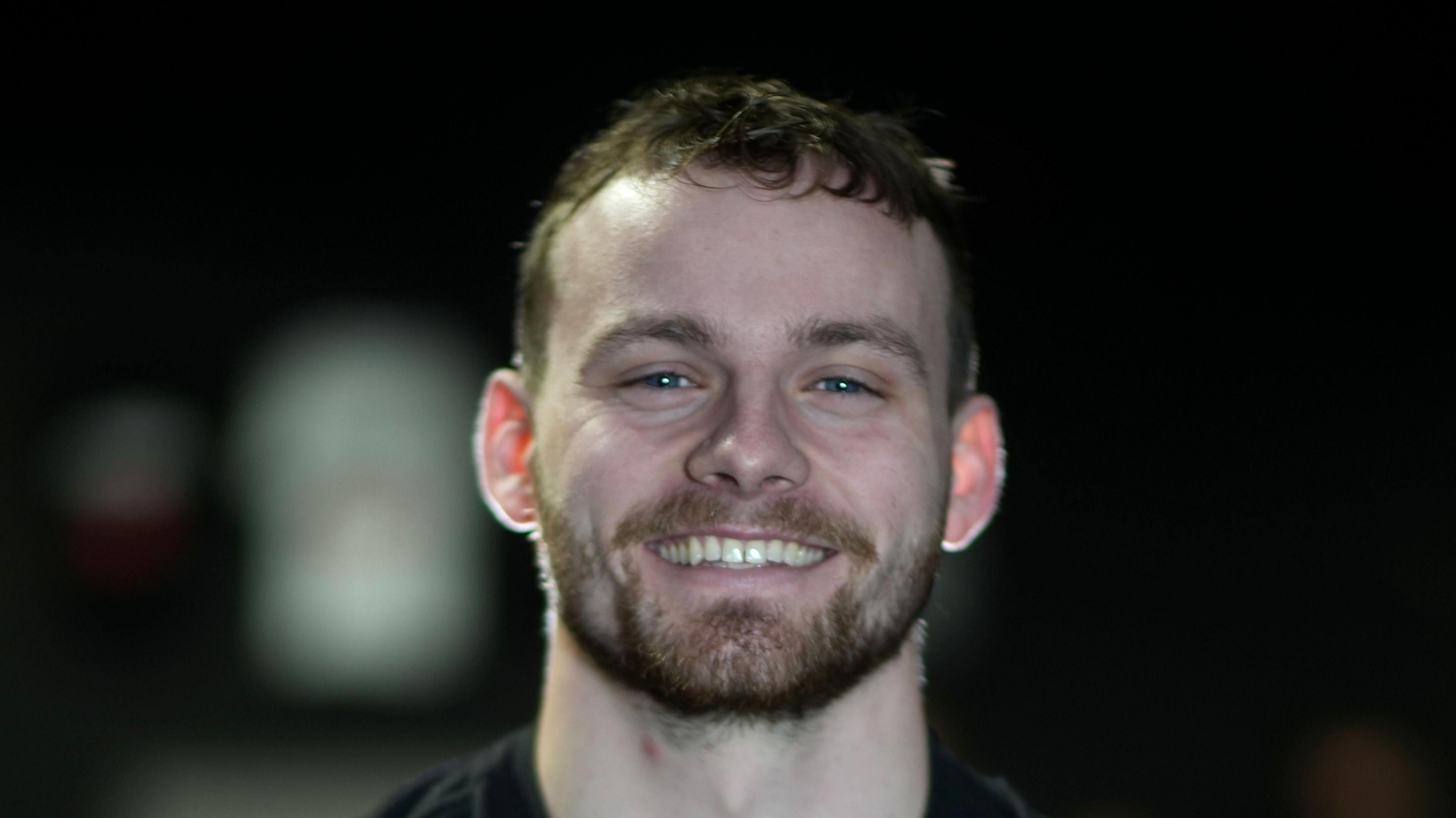 Callum Foster with short dark hair and a beard smiles at the camera. He is wearing a black top and the background is dark and blurred.