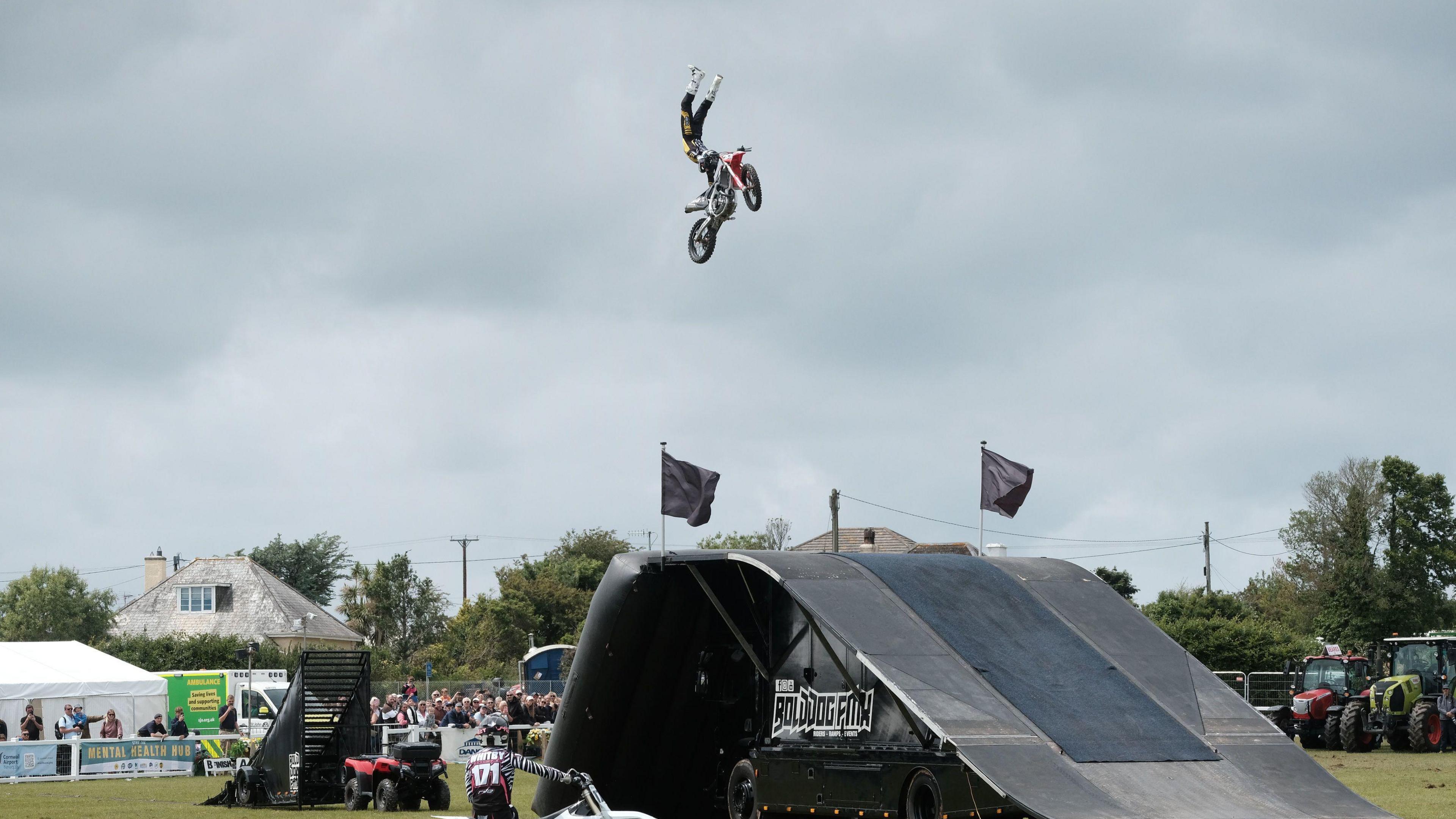 Dirt bike jumping takes place at the Royal Cornwall Show. A man and his bike are high in the air above a ramp. He is holding onto the handlebars, but his legs and feet are high above his head. Spectators can be seen in the background. 