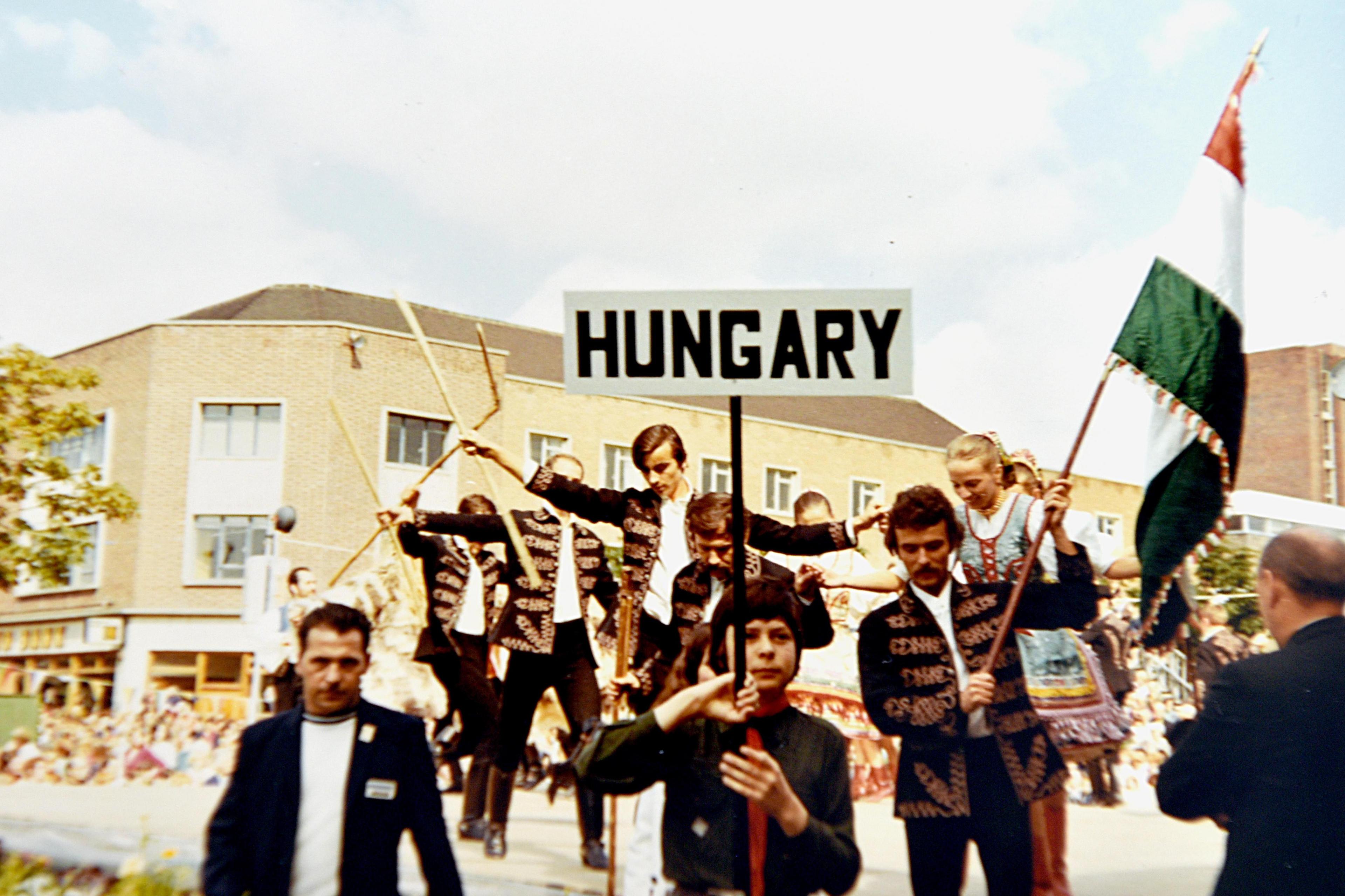 A group of men in black jackets and shirts with gold tassels holding a sign with the word Hungary on it and the flag of that country.