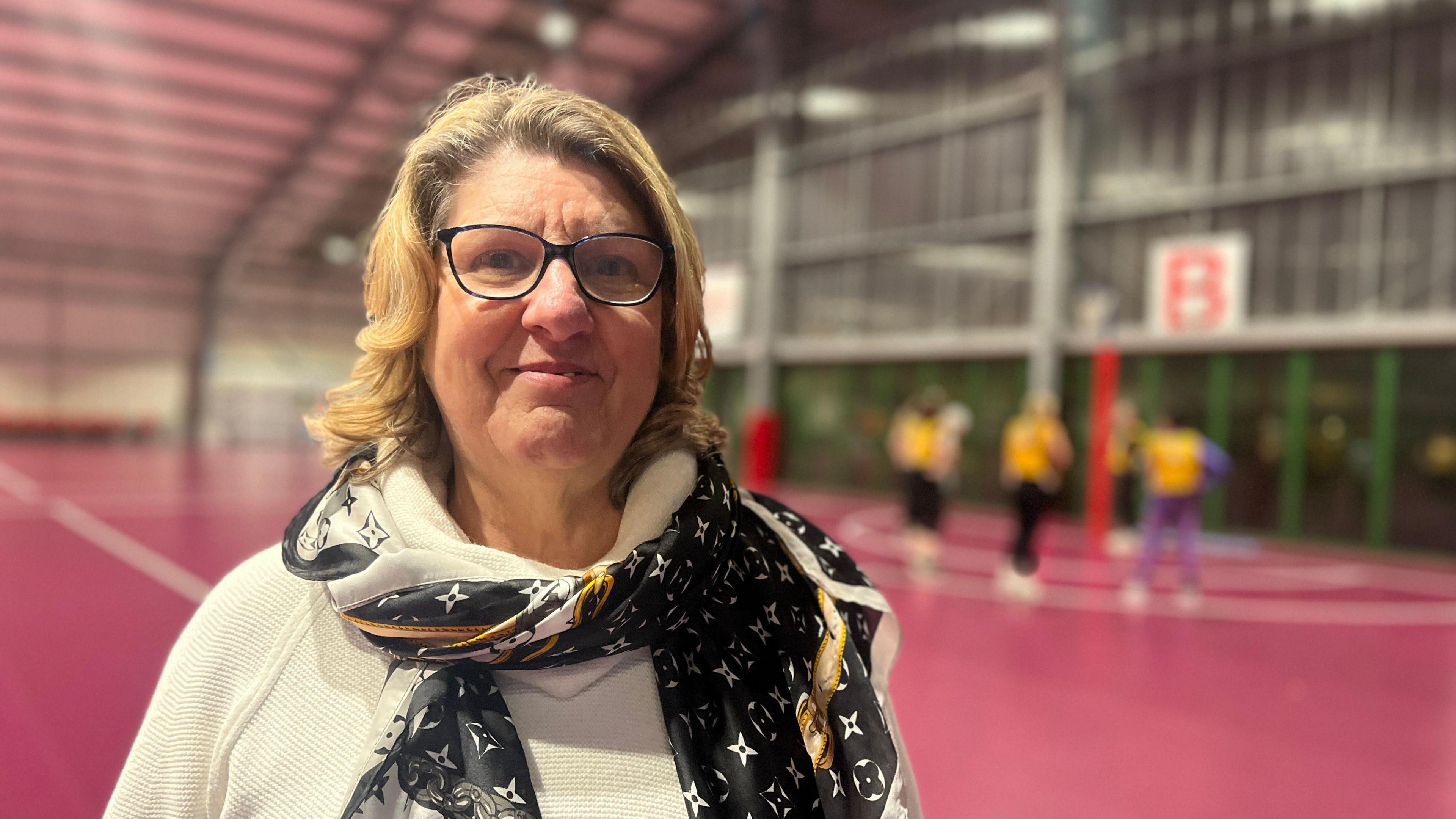 Linda looks at the camera as players practice shooting into the netball hoop behind her. She is wearing a white turtleneck jumpers with a black patterned scarf. She has light brown hair which is shoulder length. She is also wearing glasses with a dark rim.